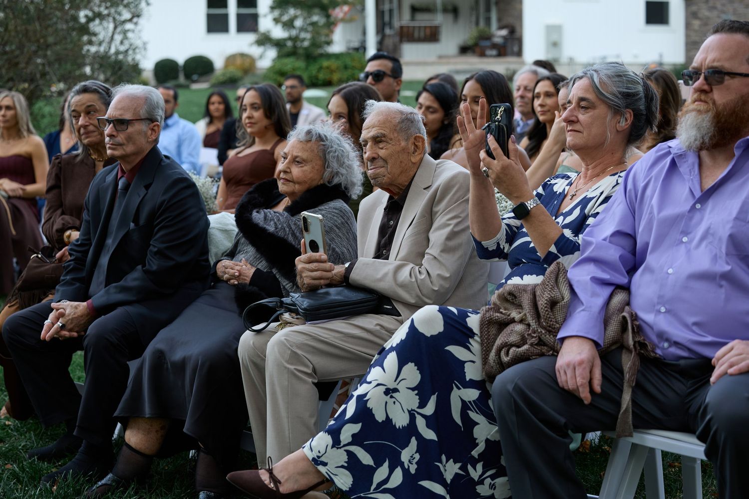 Wedding guests seated on folding chairs on the lawn clap and smile as they watch an outdoor ceremony unfold.
