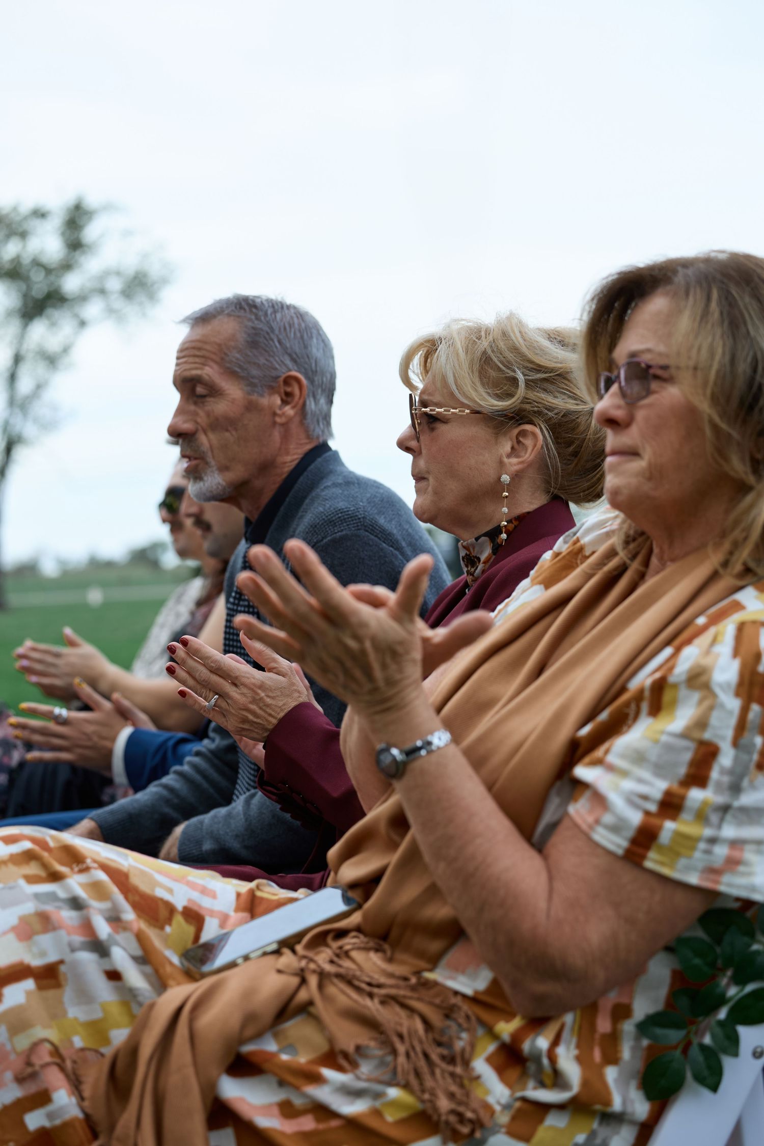 Three wedding guests applaud from their seats at an outdoor ceremony, with a woman in a colorful wrap in the foreground.