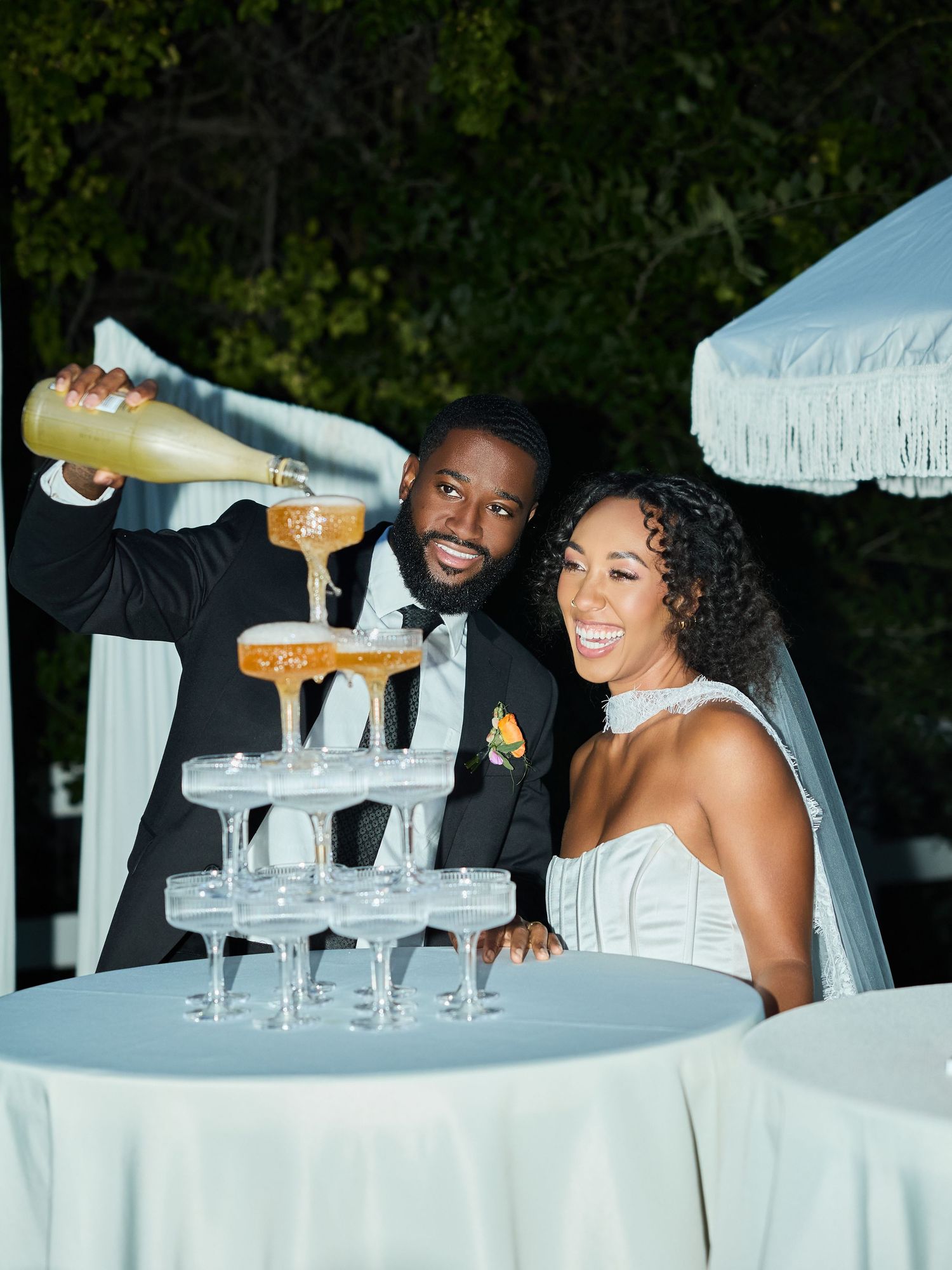 A smiling groom pours champagne into a coupe glass tower while his laughing bride in a white gown looks on at an outdoor wedding reception.
