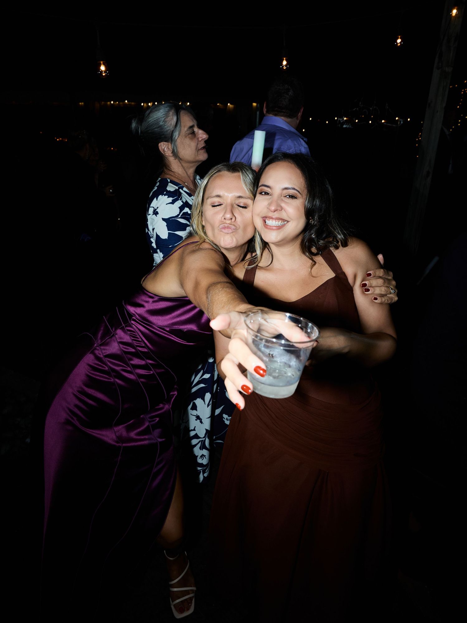 Two smiling women in satin dresses hug and pose with a drink at a lively evening wedding reception.