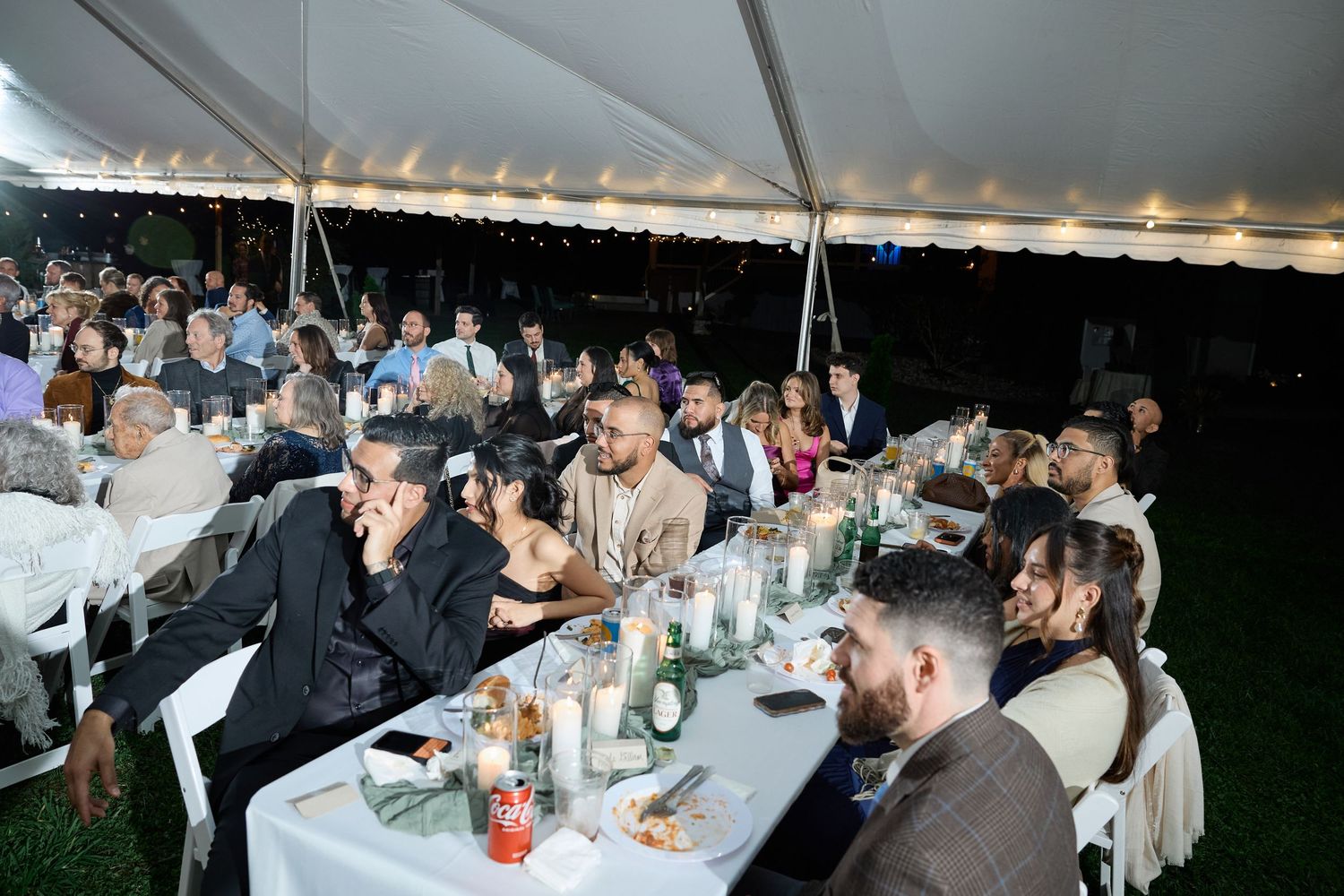 Wedding guests gather at candlelit tables under a lit tent while enjoying dinner and listening to speeches at an evening reception.