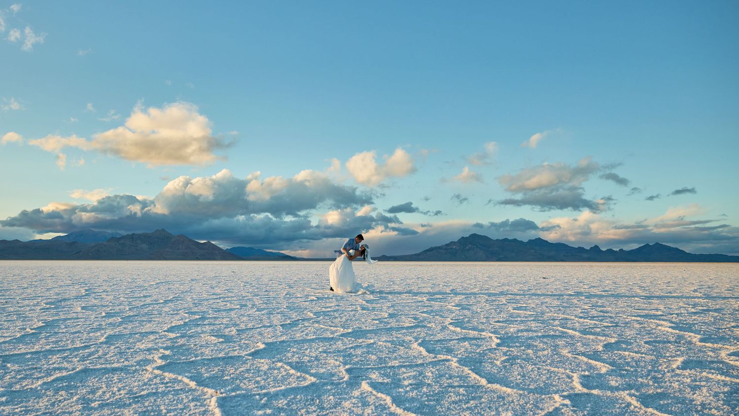 Bride in a white gown and veil poses alone on a wide salt flat with puffy clouds and mountain silhouettes in the background.