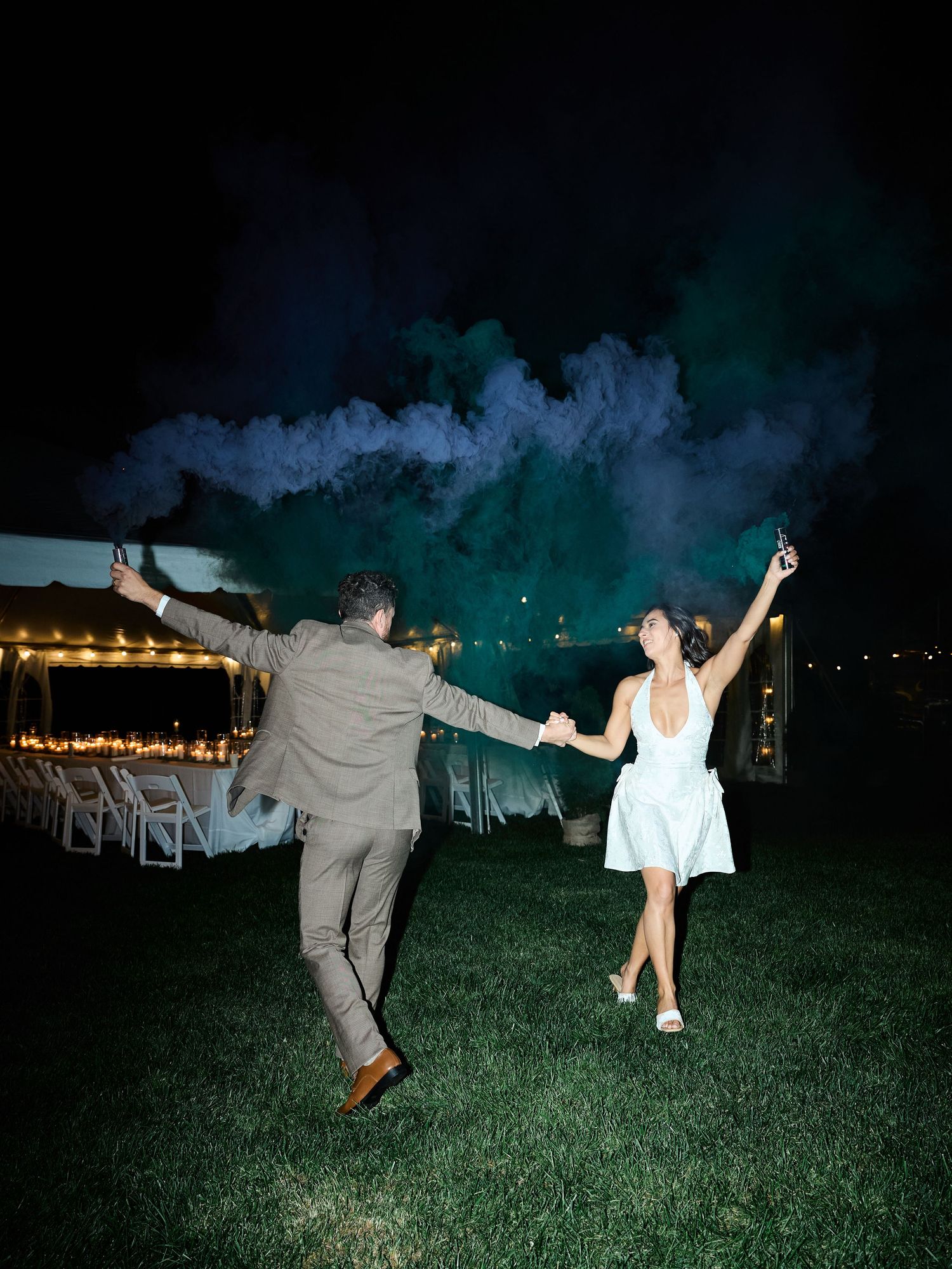 A couple holds hands and dances on a lawn at night as teal smoke bombs billow during a wedding celebration.
