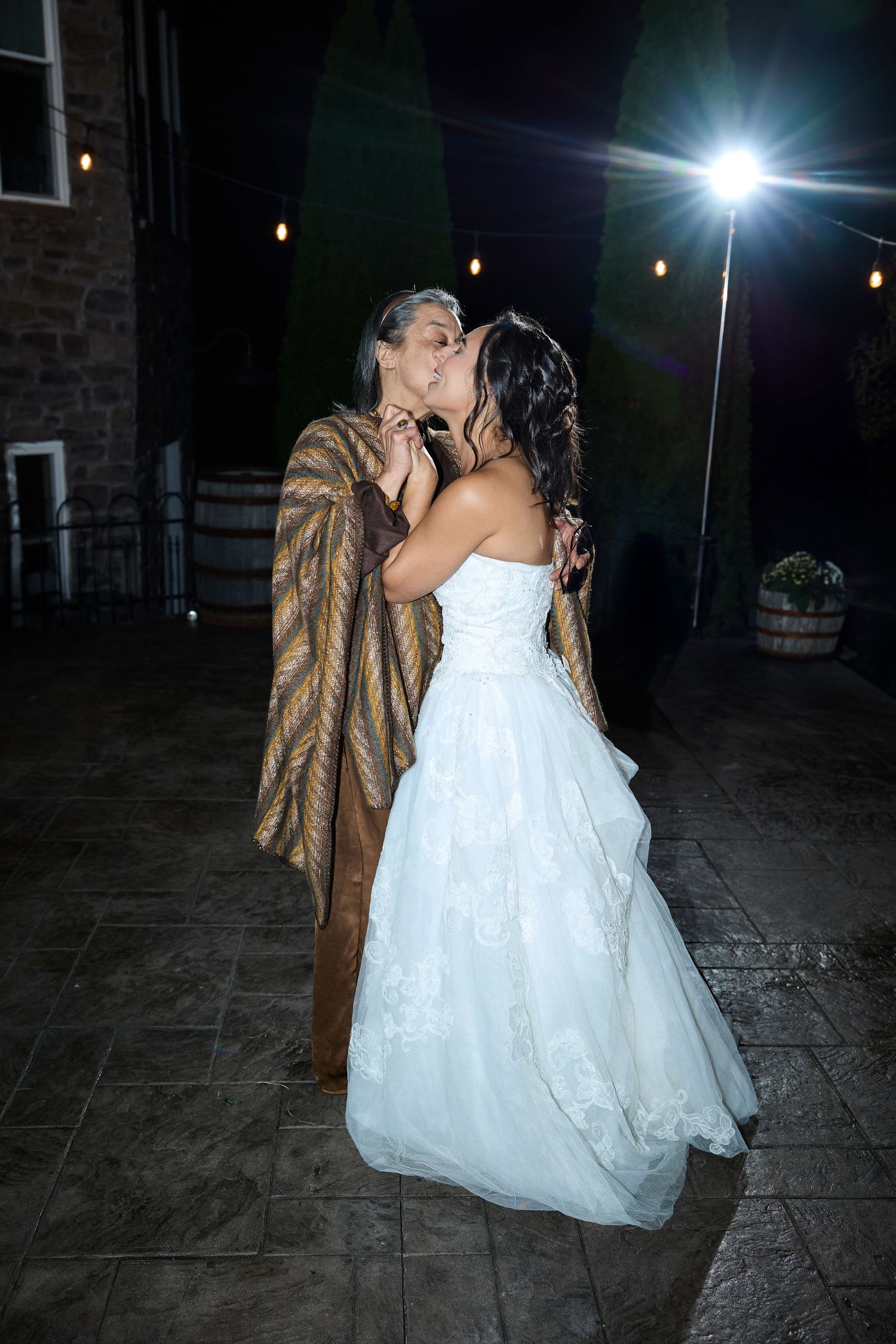 A bride in a strapless white lace gown kisses a woman wrapped in a gold and brown shawl under glowing string lights.