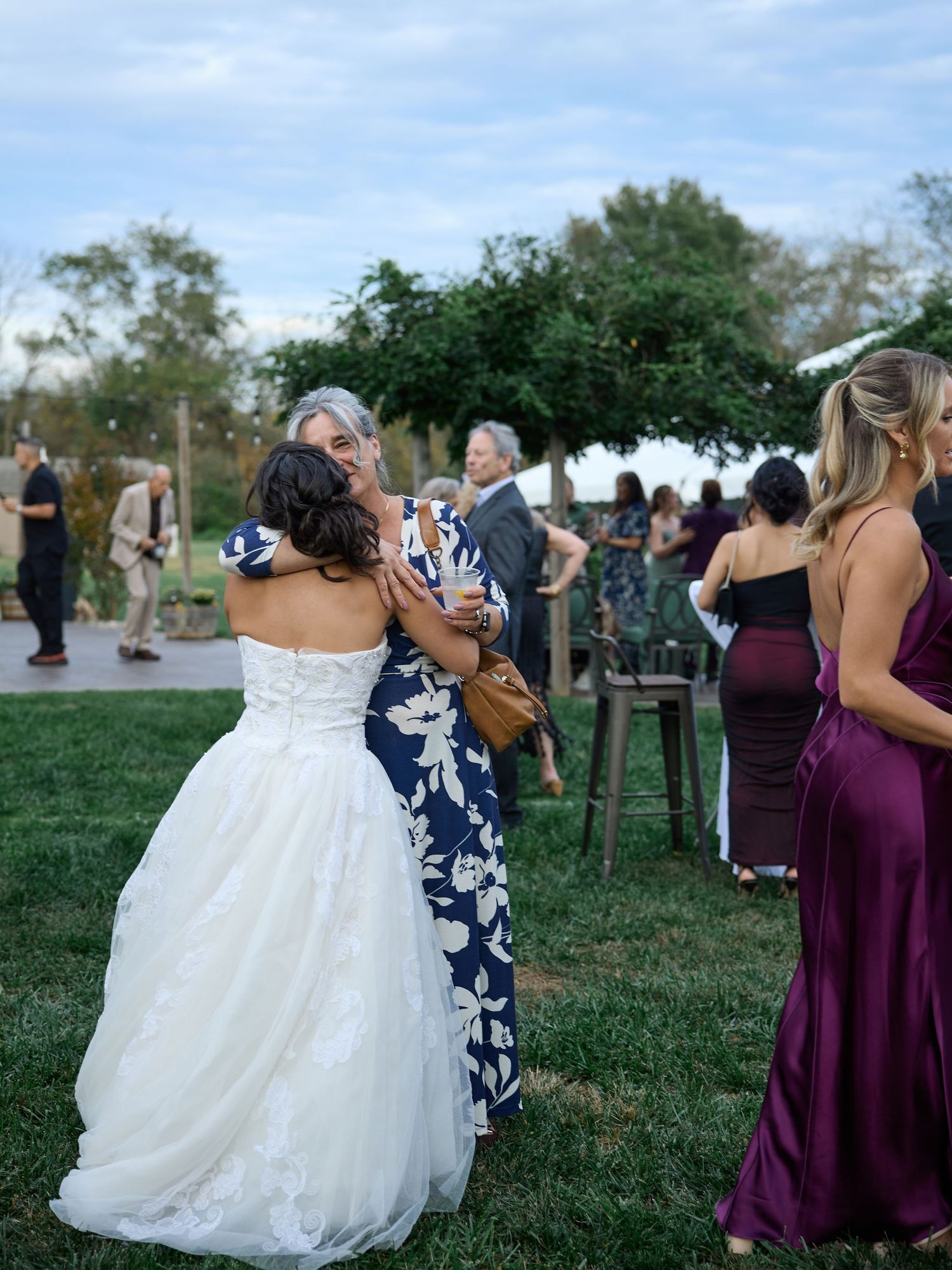 A bride in a strapless white gown hugs a guest in a floral dress on the lawn as bridesmaids in plum dresses look on.