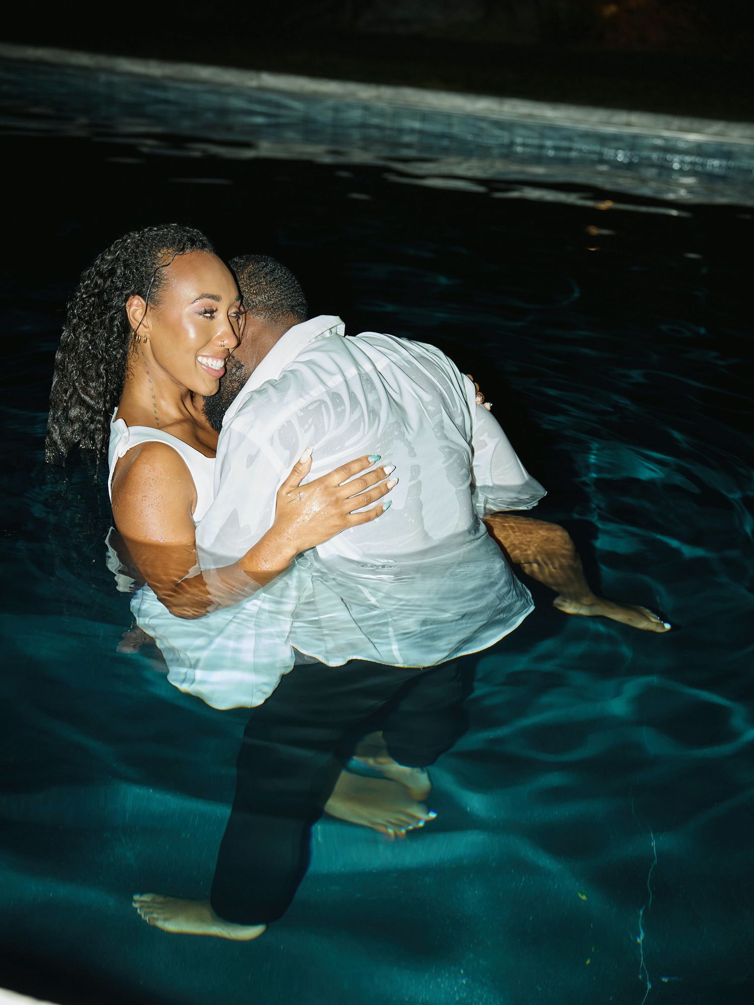 A smiling woman in white is carried by a man in a wet white shirt through a dark pool at night.