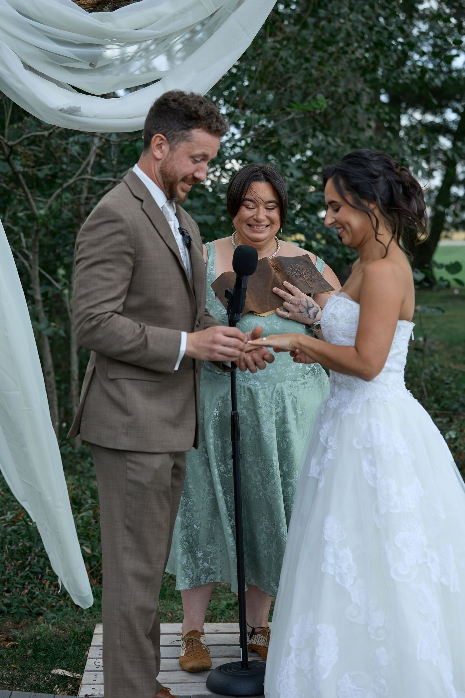 A smiling groom in a tan suit exchanges rings with the bride while the officiant in a green dress looks on at the ceremony.