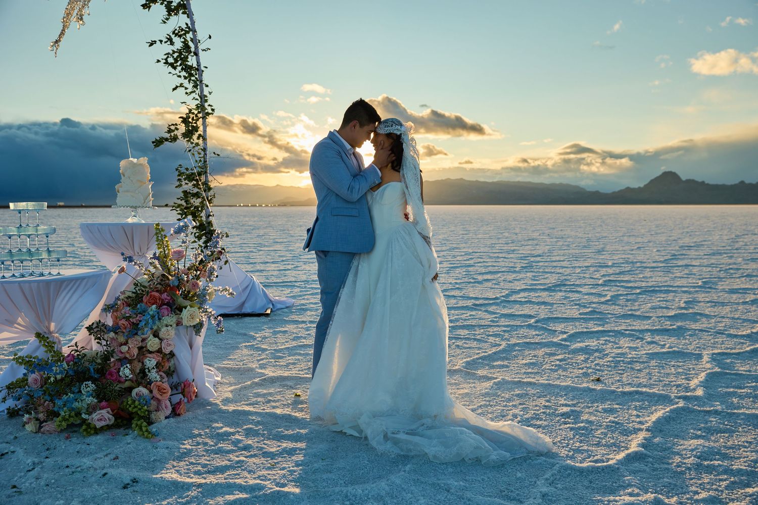 Bride and groom kiss at sunset beside a floral arch and champagne tower on a cracked white salt flat wedding venue.