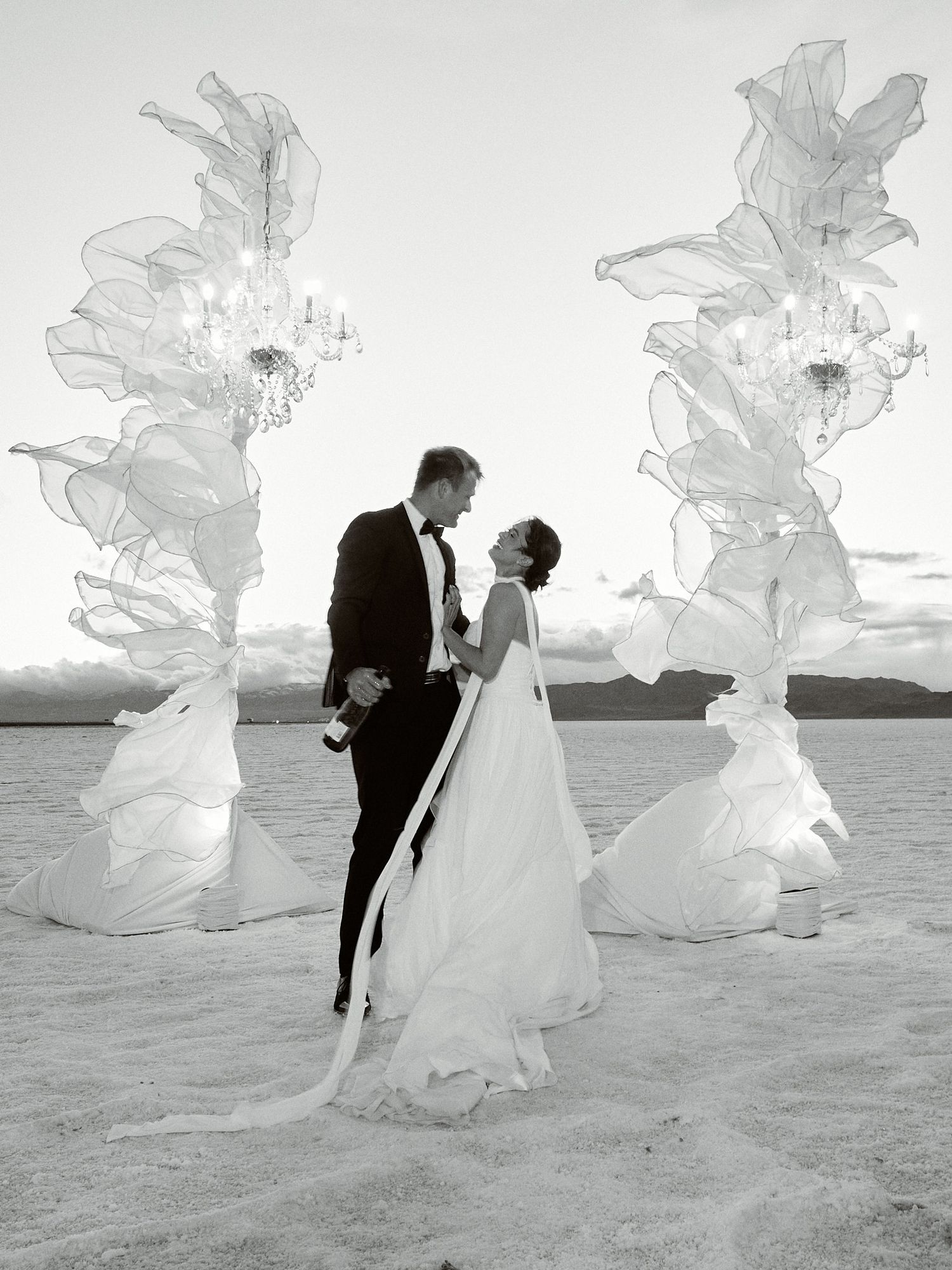 Black and white image of a bride and groom embracing between two tall sheer fabric chandelier installations on a flat desert landscape.