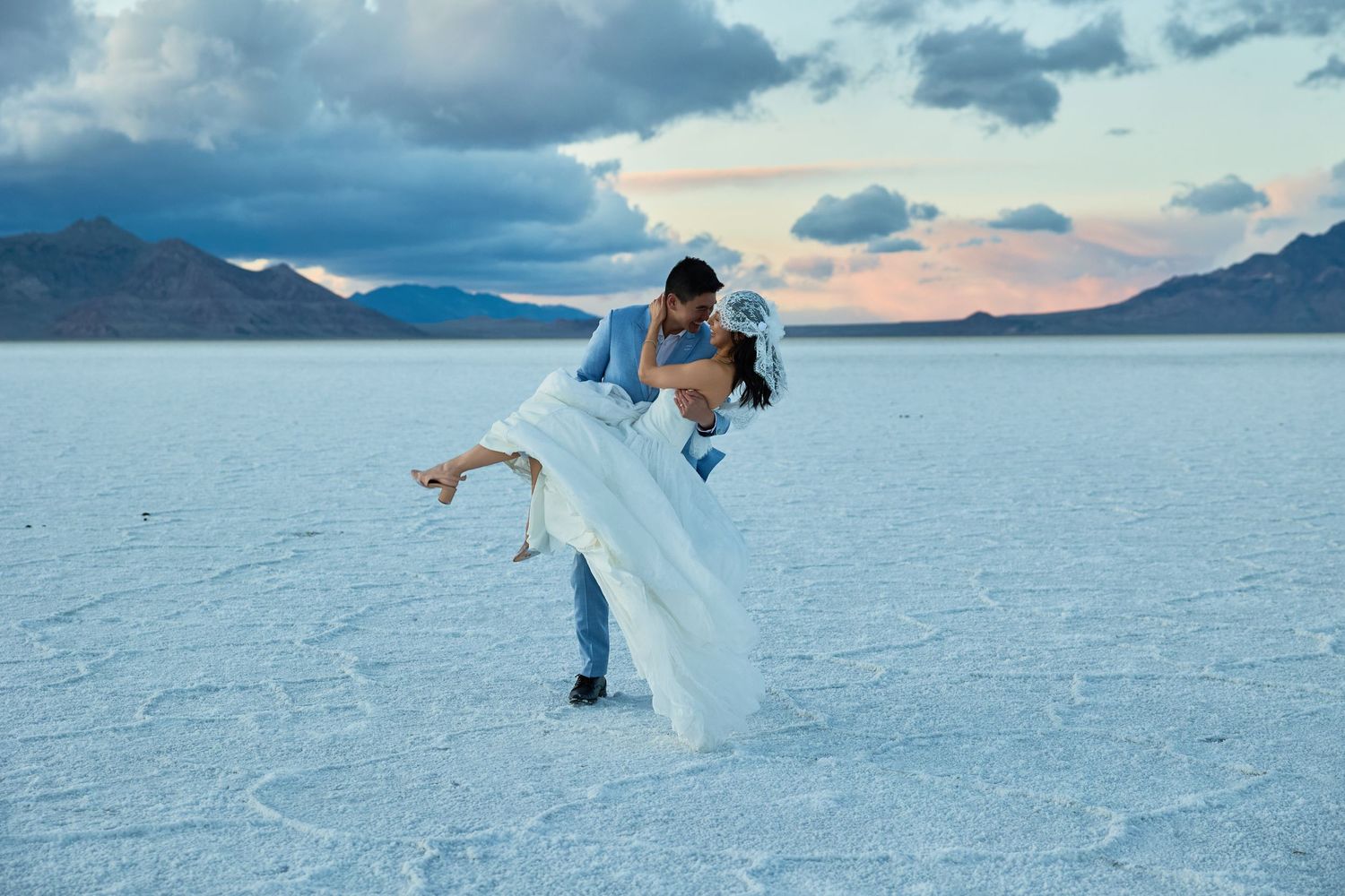 Groom lifts and kisses his bride in a lace veil on a cracked salt flat with mountains and colorful clouds behind.