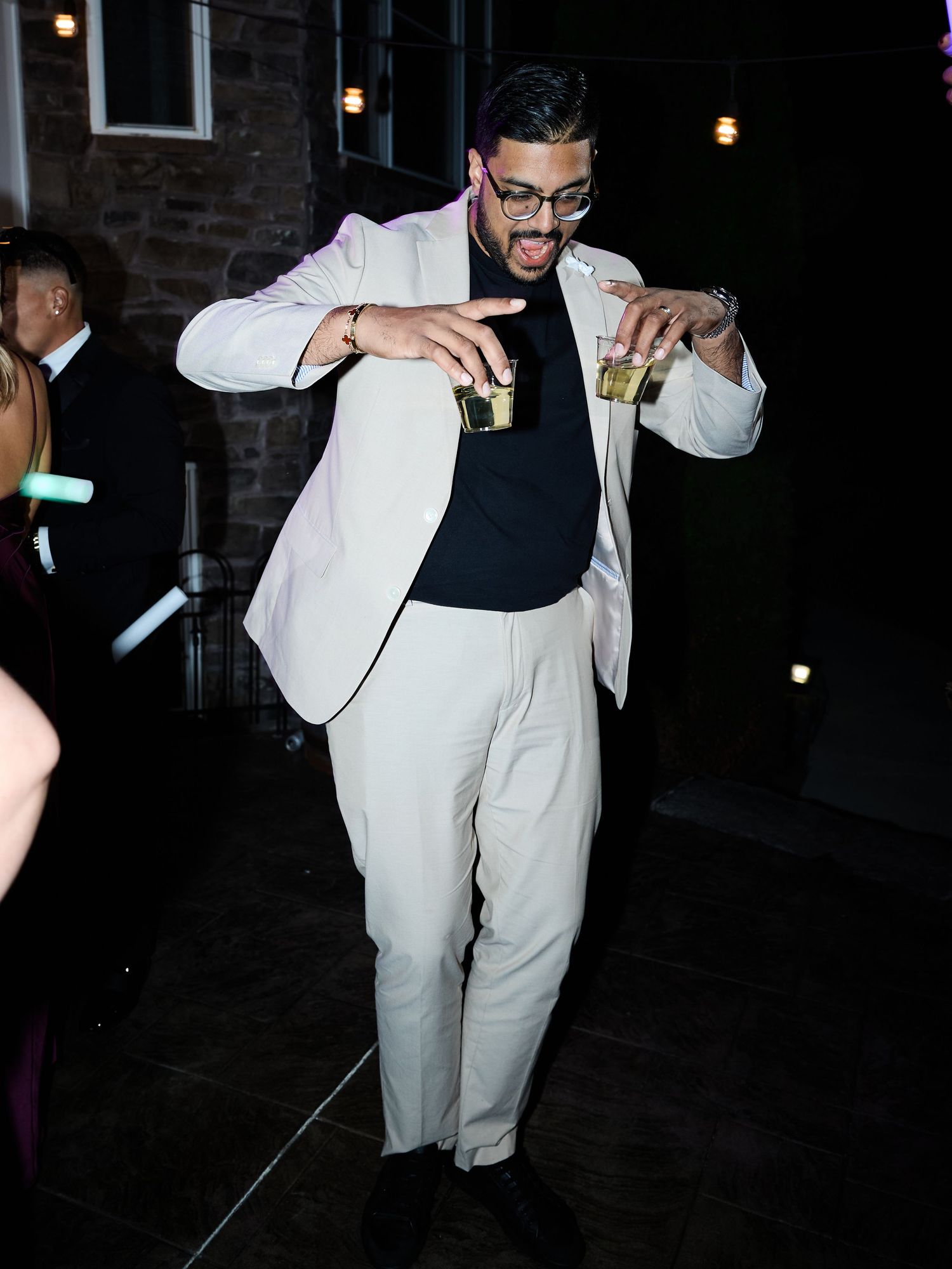 A man in glasses and a cream suit dances at a night wedding reception while holding two small drinks.