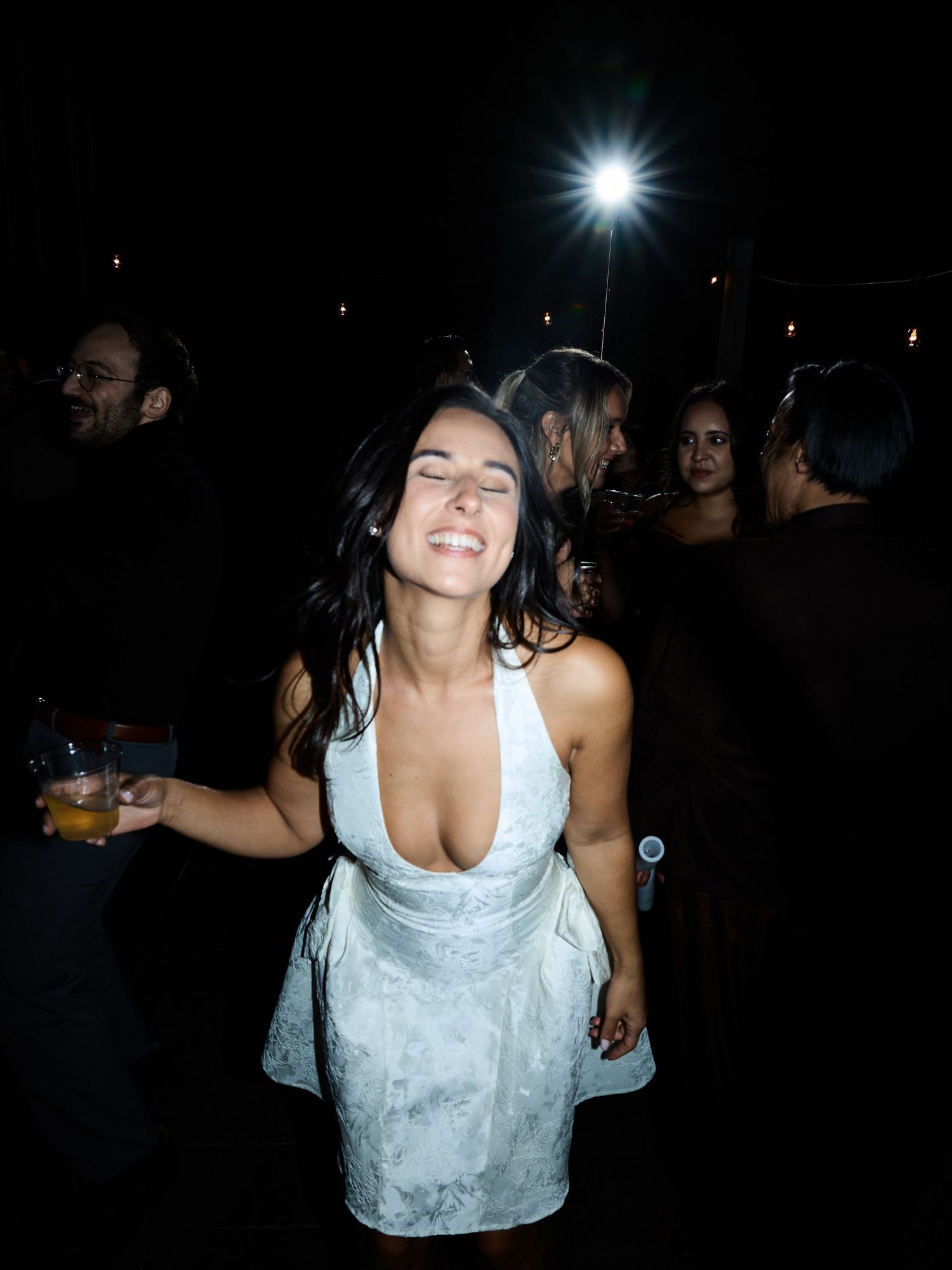 A laughing woman in a white satin dress holds a drink and dances joyfully at an evening wedding party.