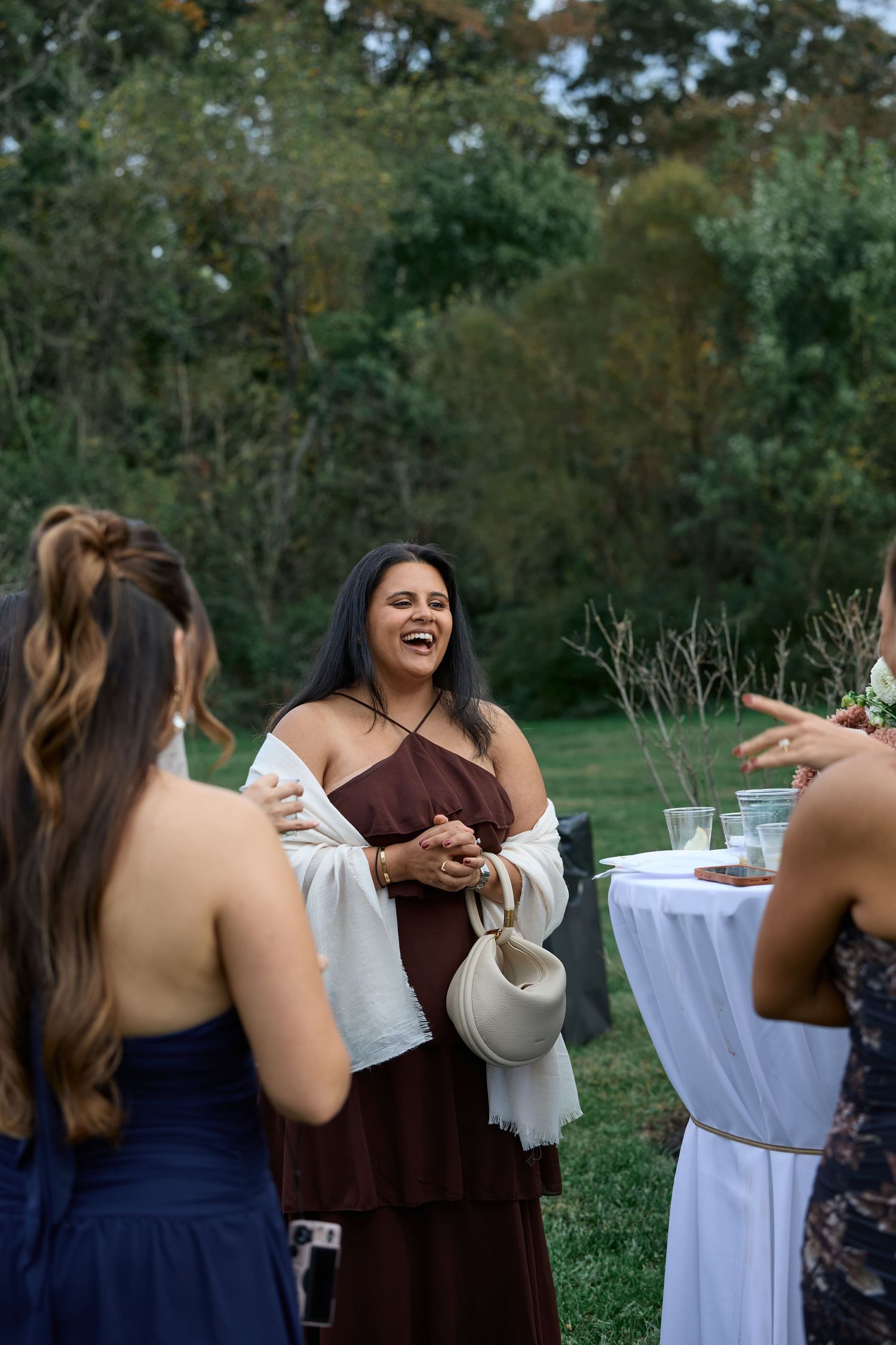 A smiling woman in a burgundy dress and white shawl laughs while talking with other guests at an outdoor garden celebration.