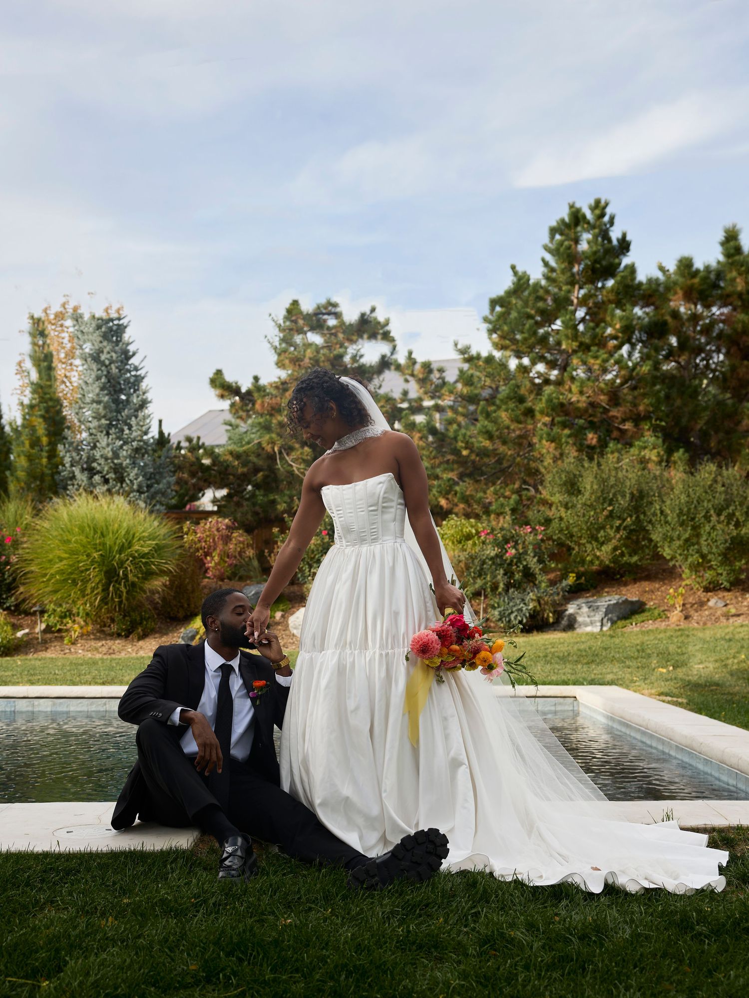 A bride in a strapless white gown holds a colorful bouquet while her groom in a black suit sits by a pool on the lawn.