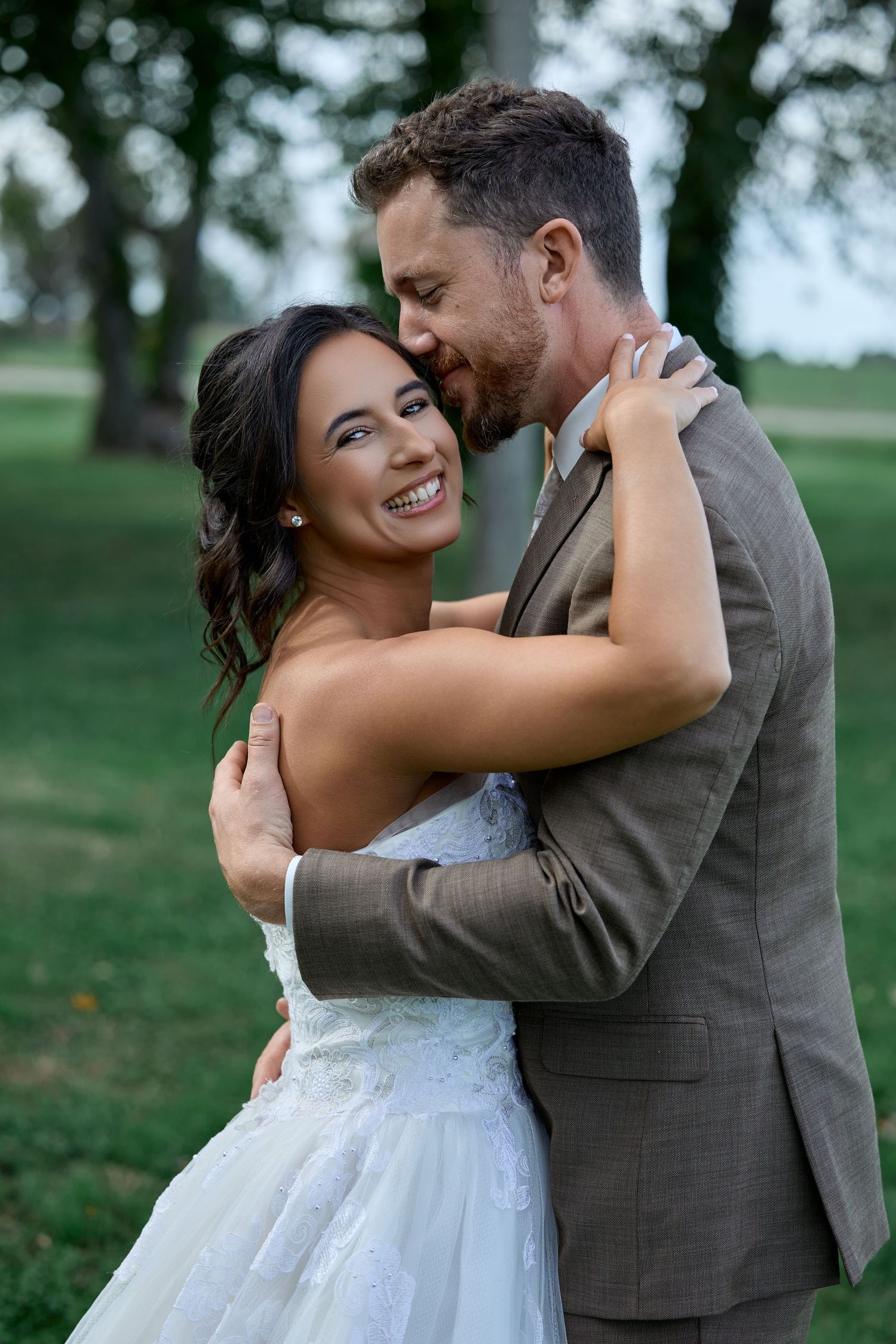 Bride in a white lace gown smiles warmly as her groom in a brown suit kisses her cheek during an outdoor embrace.