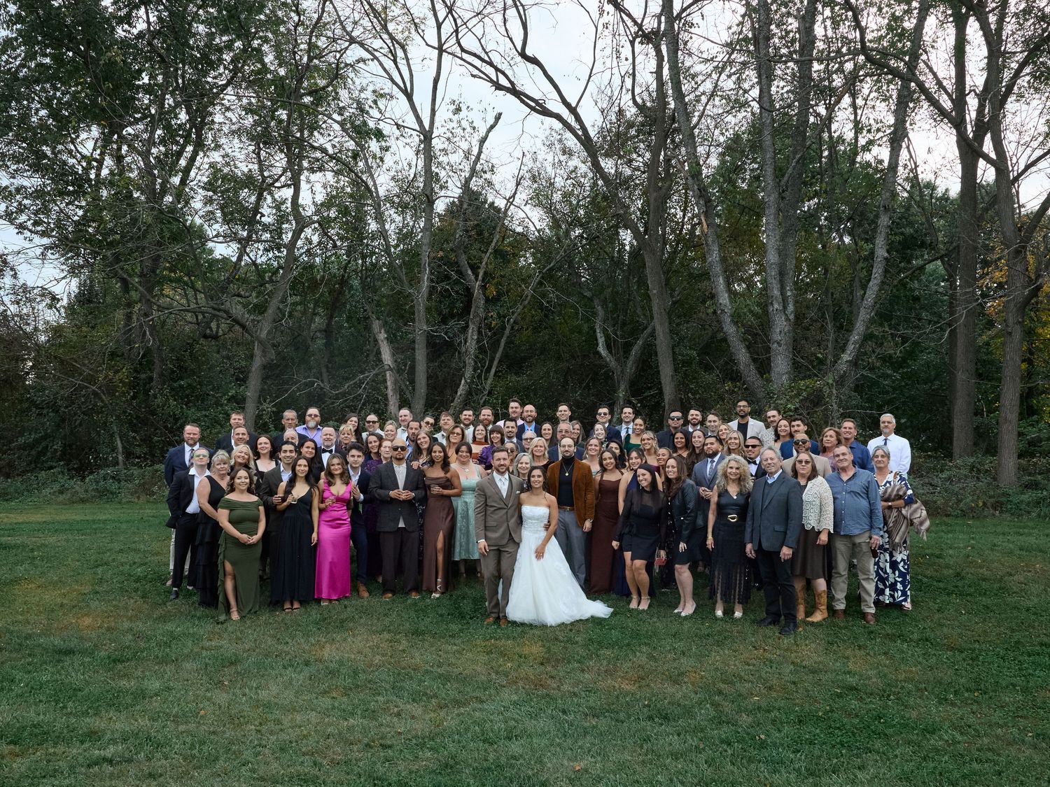 A full wedding party poses together with the bride in white on a lawn framed by tall trees in autumn.