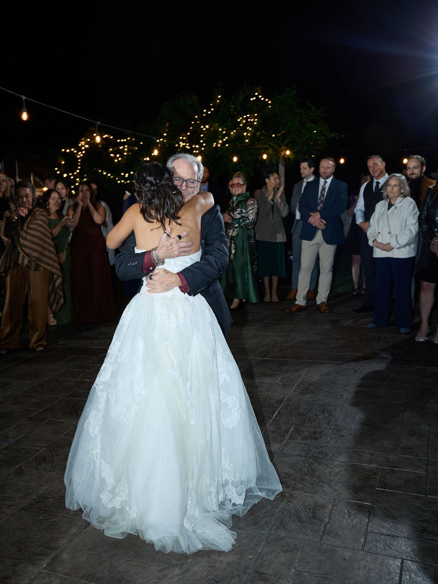 A bride in a white lace wedding gown shares an embrace with an older man while guests look on at a night reception.