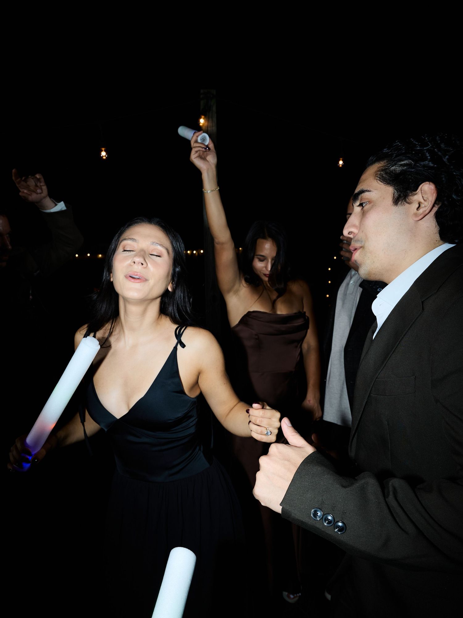 A woman in a black dress dances with a glow stick while guests celebrate around her at a night wedding.