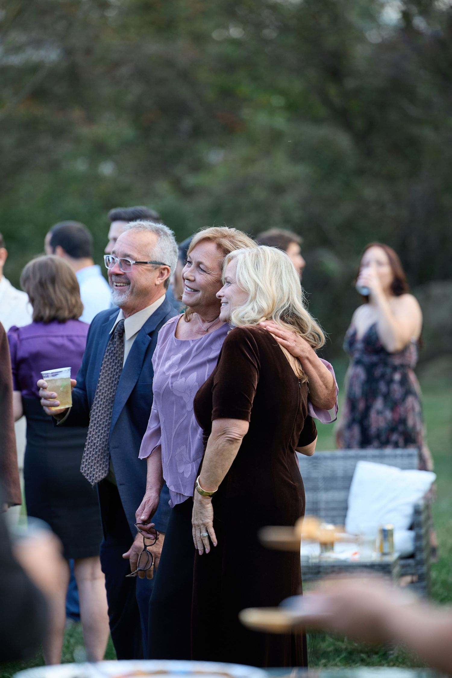 Smiling older guests stand arm in arm at an outdoor wedding reception, enjoying drinks as other guests mingle behind them.