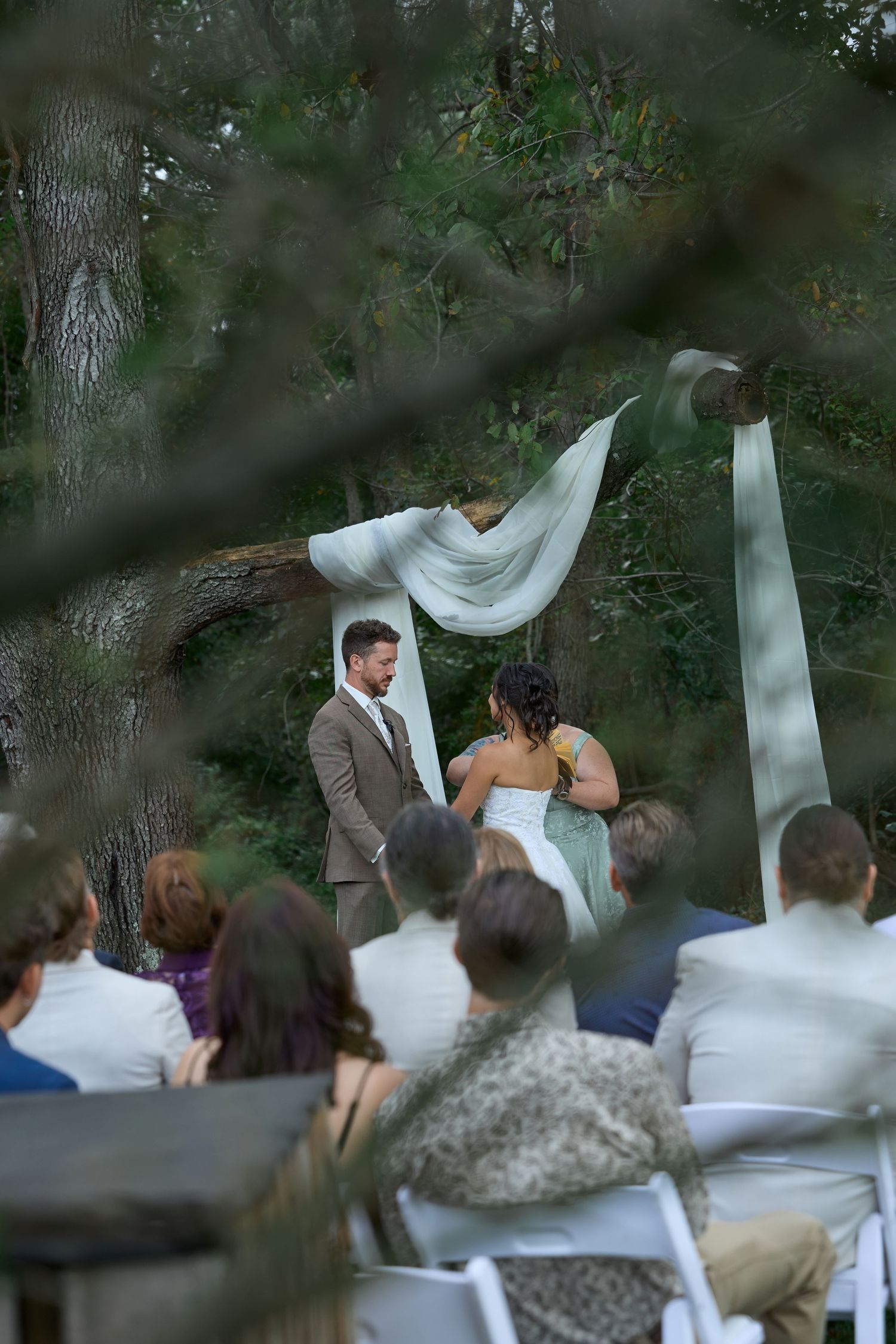 A groom and bride stand together beneath a draped fabric arch among trees as guests watch the ceremony from white chairs.