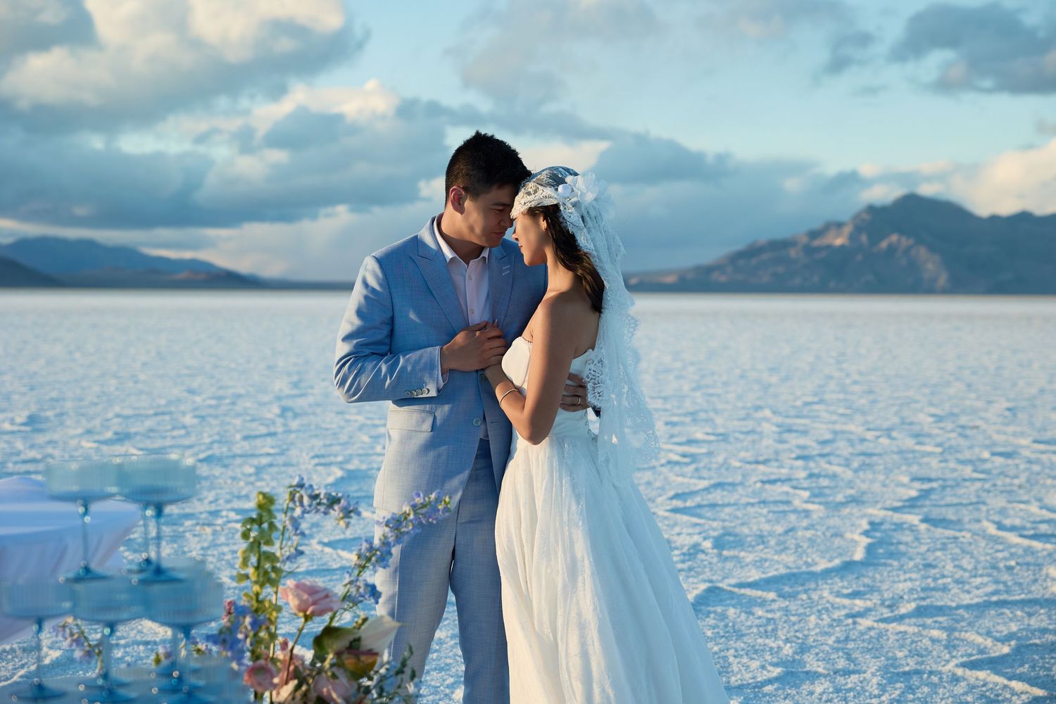 Bride in a white gown and veil embraces her groom in a light blue suit on a reflective salt flat at dusk.