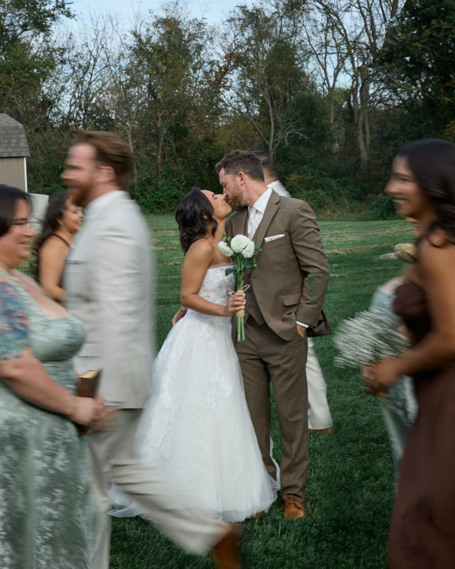 Bride and groom kiss on a lawn as blurred wedding guests in motion walk past them in the foreground.