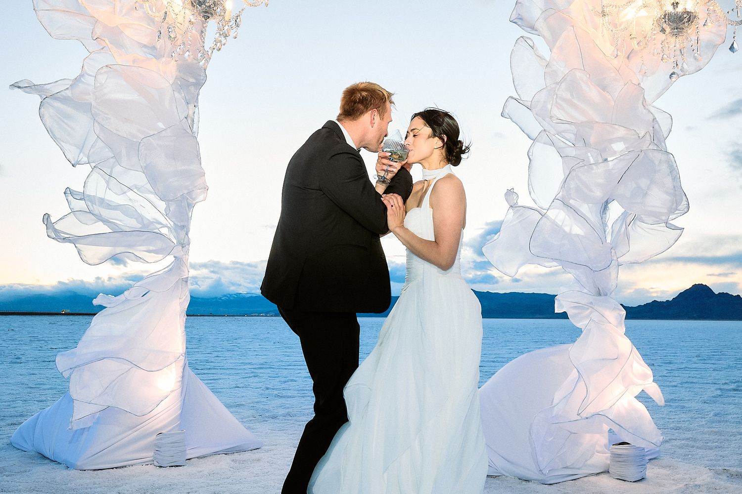 A bride and groom share a toast at sunset between two glowing sheer fabric chandelier sculptures beside a calm lake.