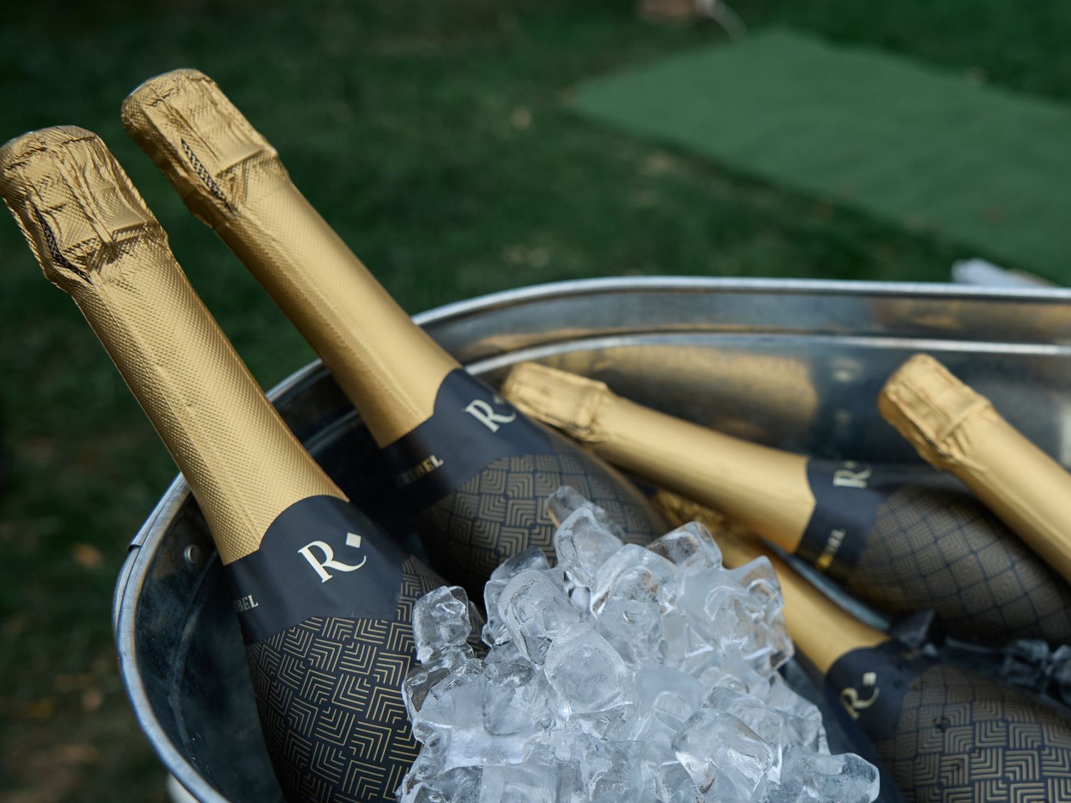 Several champagne bottles with gold foil tops chill in a metal tub of ice at an outdoor wedding celebration.