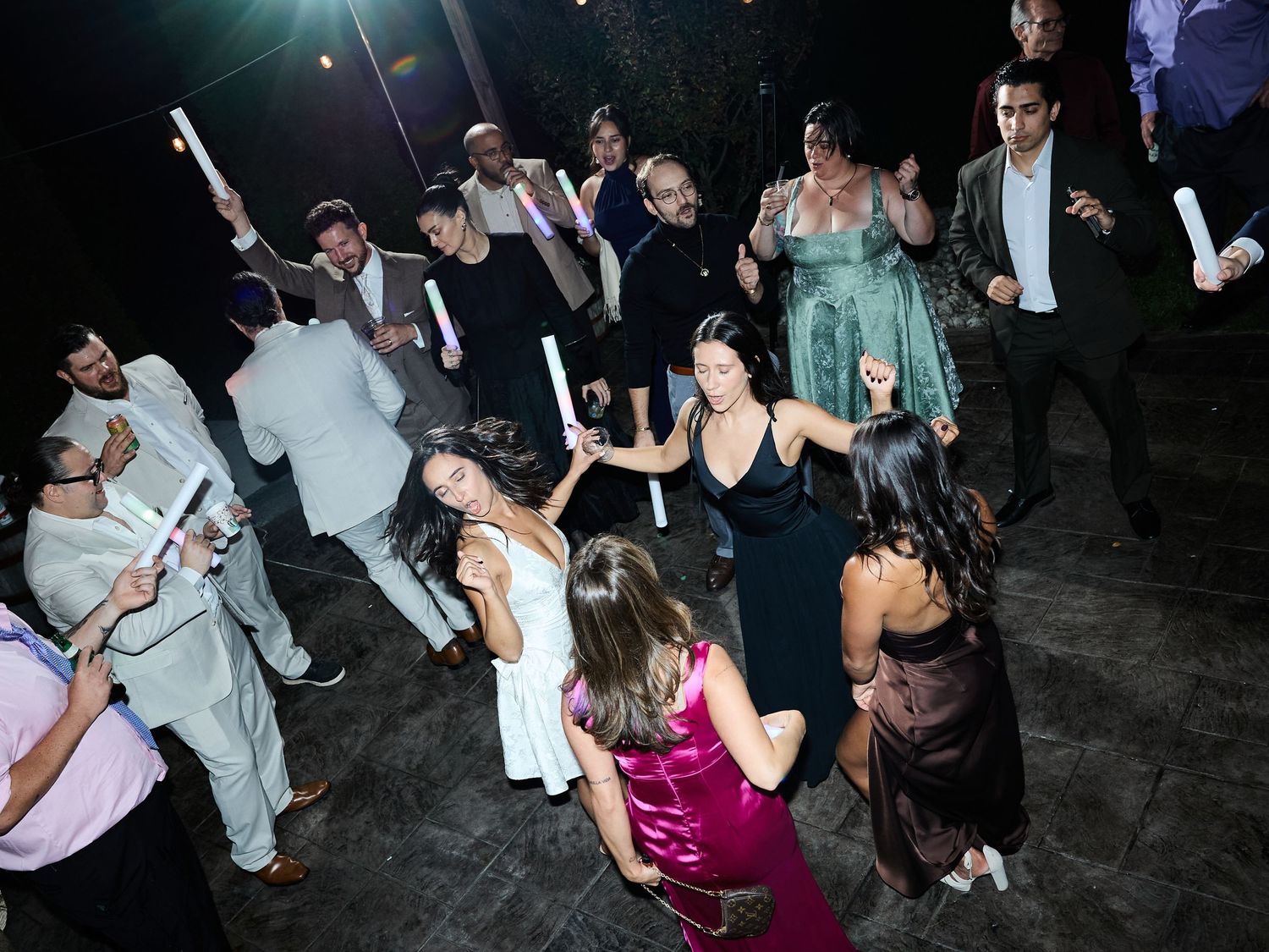 Wedding guests dance in a circle holding glowing light sticks on an outdoor dance floor at night.