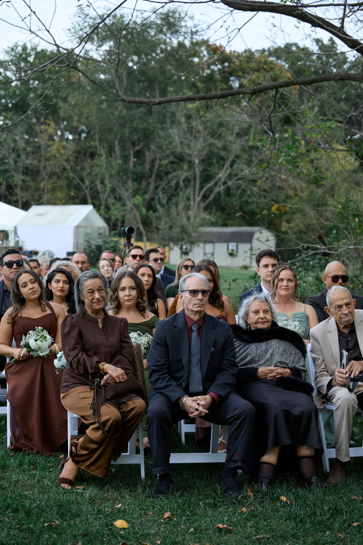 Rows of formally dressed guests, including women holding bouquets, sit quietly on white chairs at an outdoor wedding ceremony.