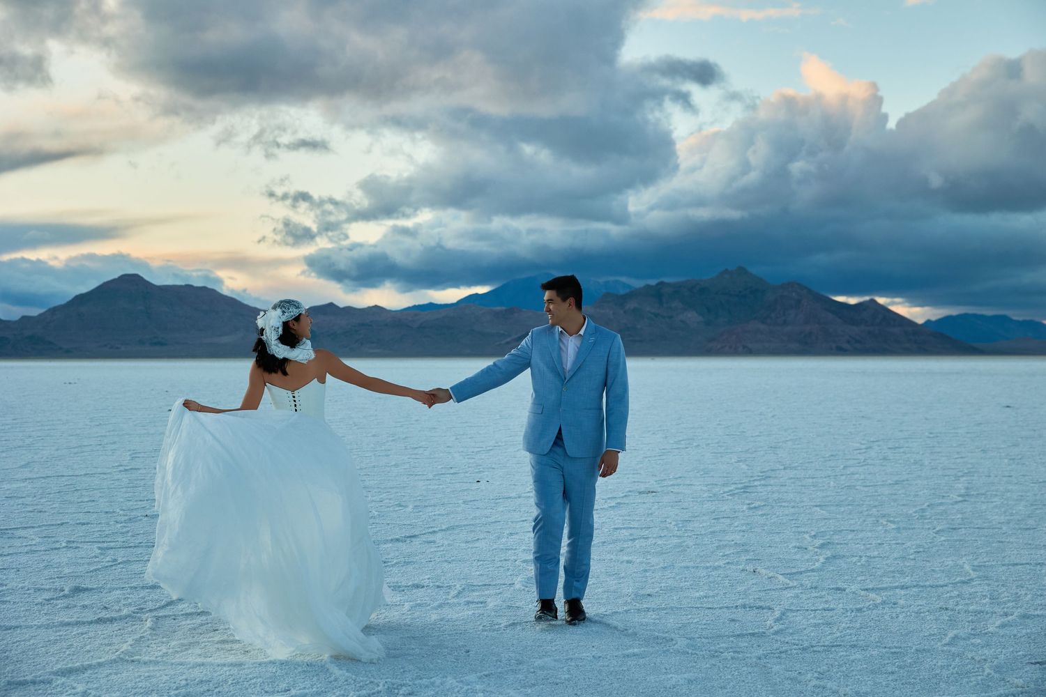 Bride and groom hold hands while facing apart on a white salt flat, with mountains and stormy clouds in the dusk sky.