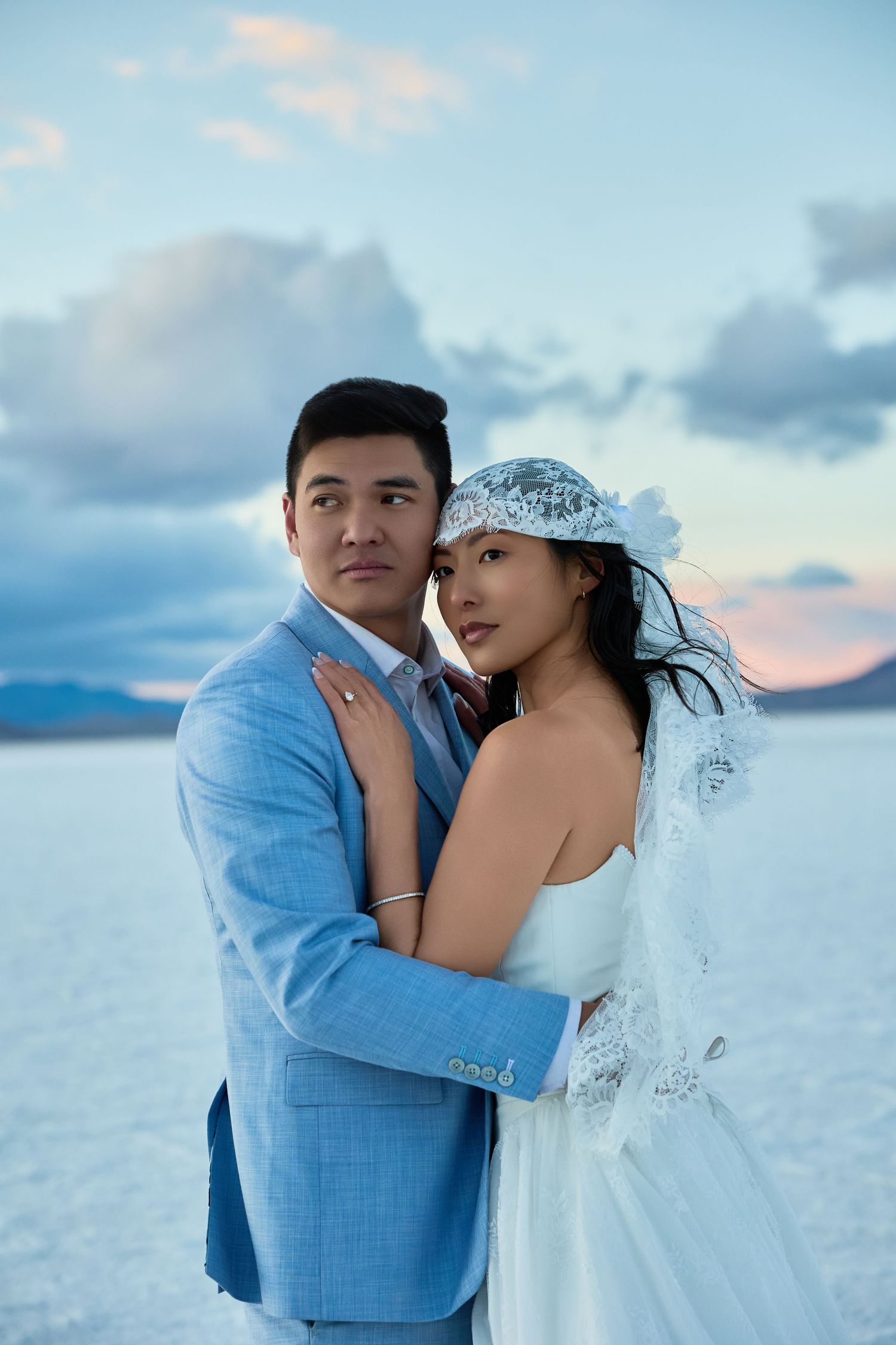 Groom in a light blue suit looks aside while the bride in a lace veil leans into him against a pastel salt flat sky.