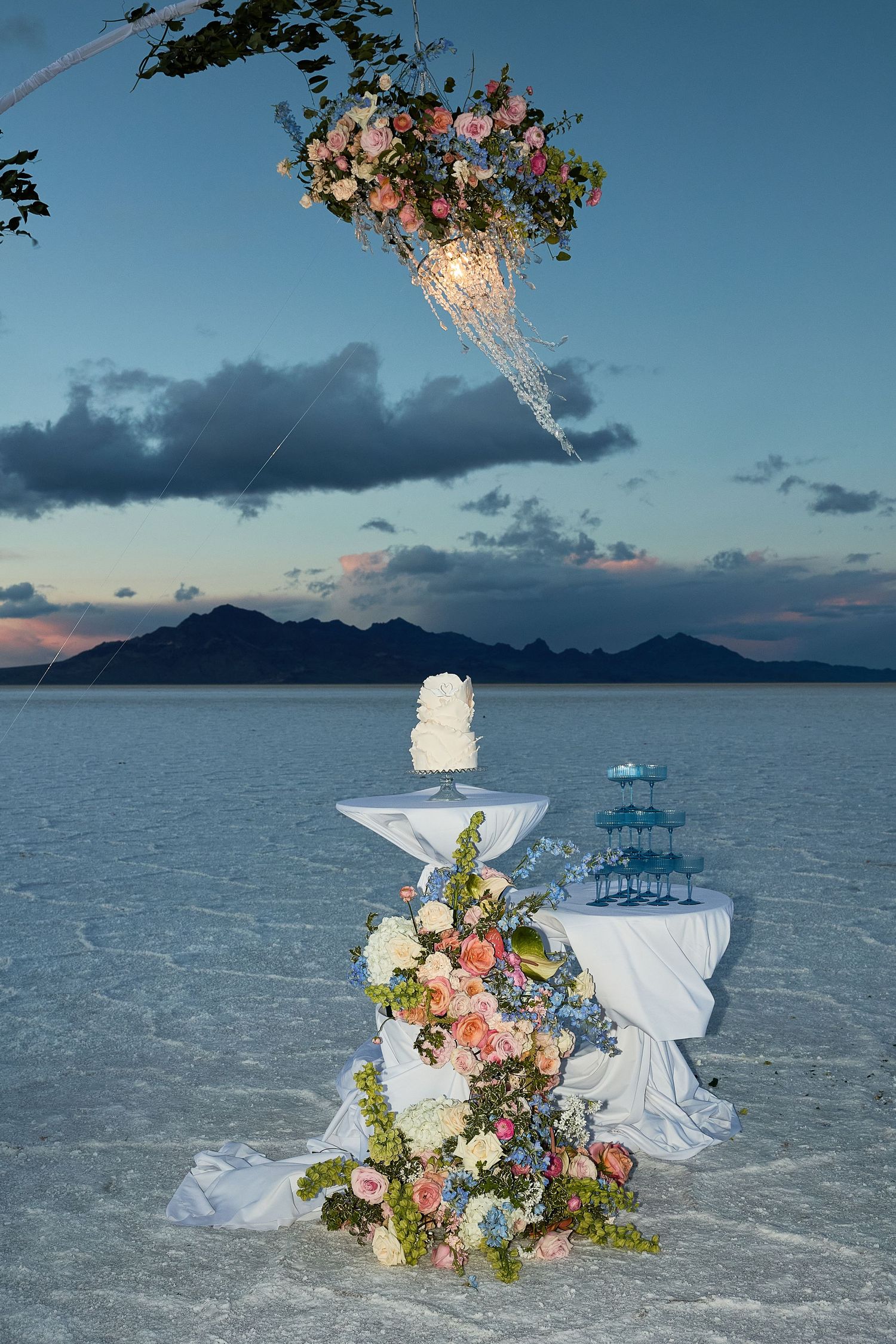 Wedding cake, floral arrangement, and champagne tower on a draped table under a hanging chandelier on a salt flat at dusk.