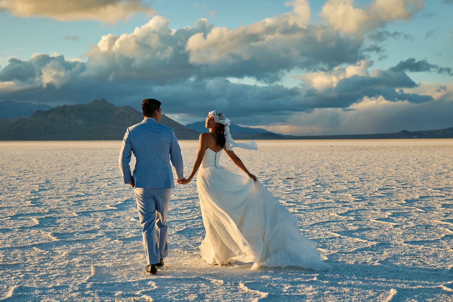 Newlywed couple walks hand in hand across cracked white salt flats toward distant mountains beneath a bright, cloud-filled sky.