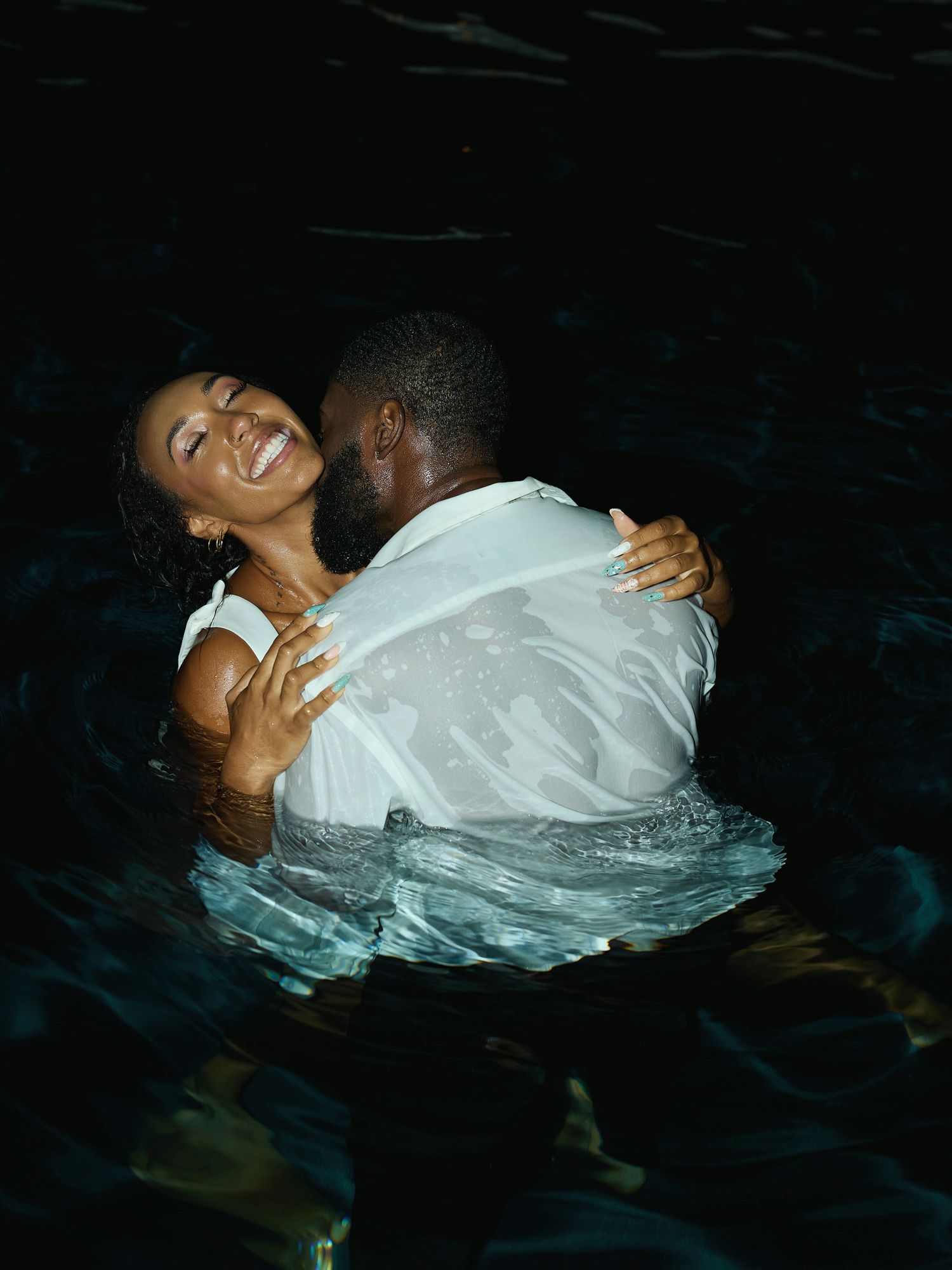 A joyful woman laughs as she embraces her partner in a wet white shirt in dark pool water at night.