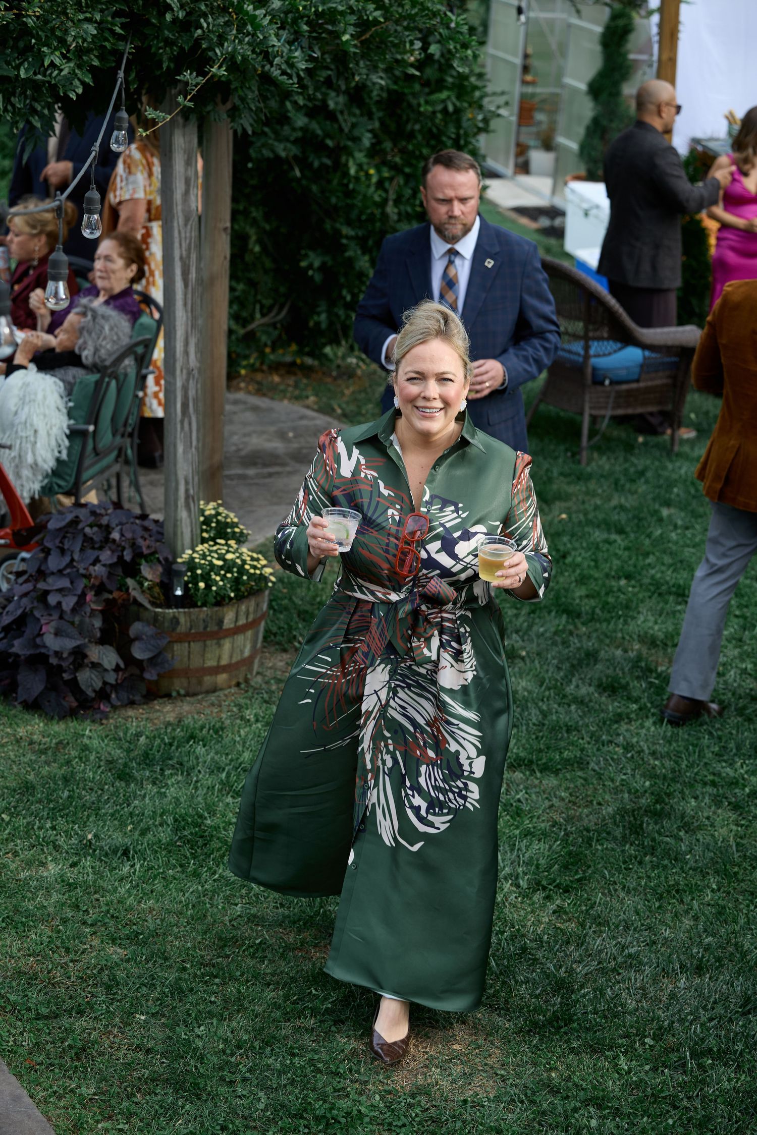 A smiling woman in a green patterned dress walks across the lawn holding two drinks while guests socialize behind her at a wedding.