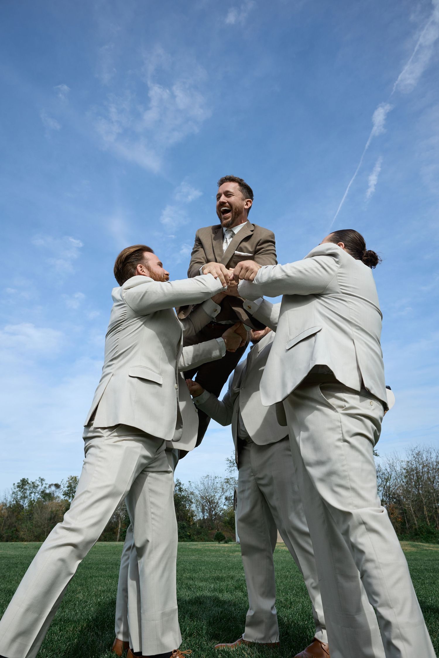 Low-angle view of groomsmen in gray suits hoisting a smiling groom skyward on a grassy field under a bright blue sky.