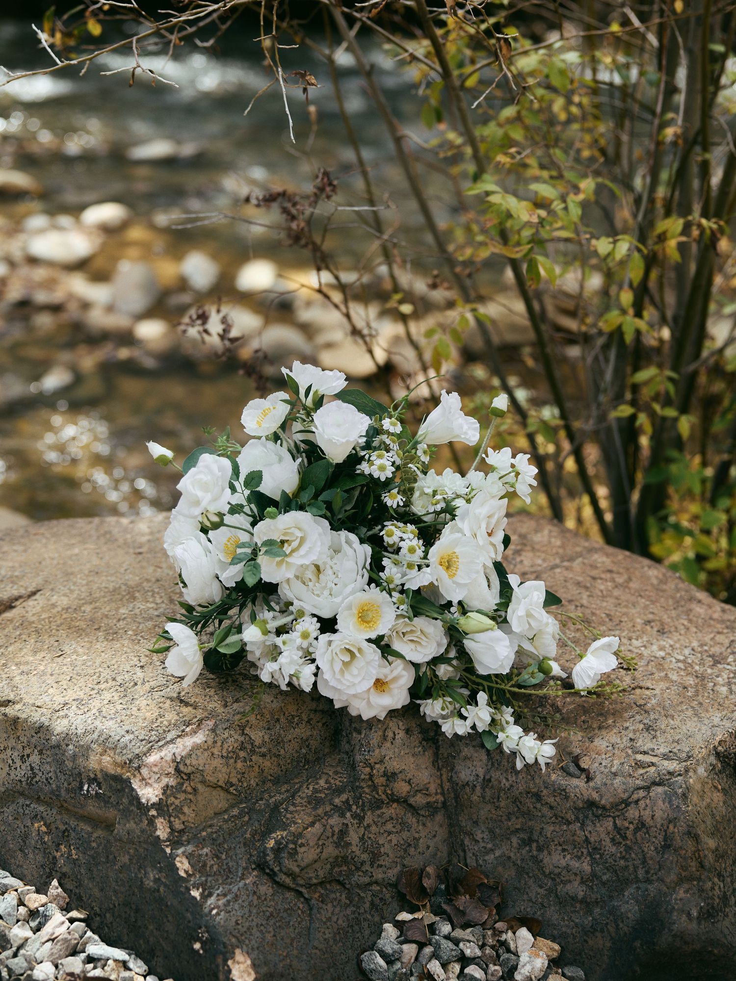 White rose and wildflower bridal bouquet resting on a large rock beside a clear rocky mountain stream.
