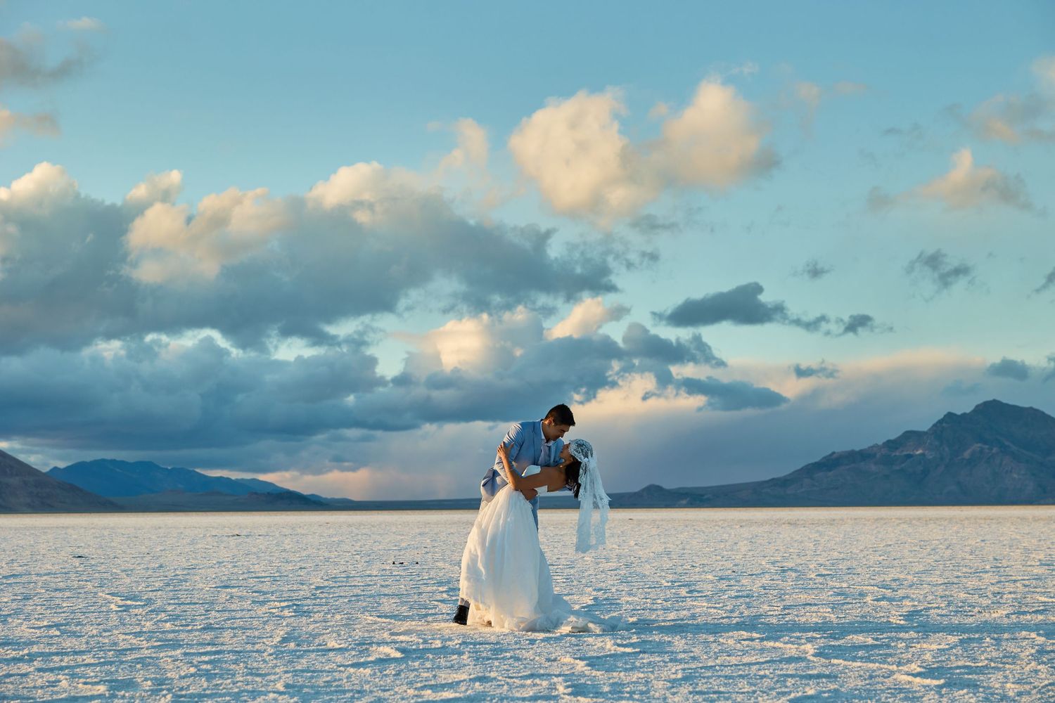 Groom dips his bride in a romantic embrace on a white salt flat beneath billowing clouds and distant mountains at dusk.