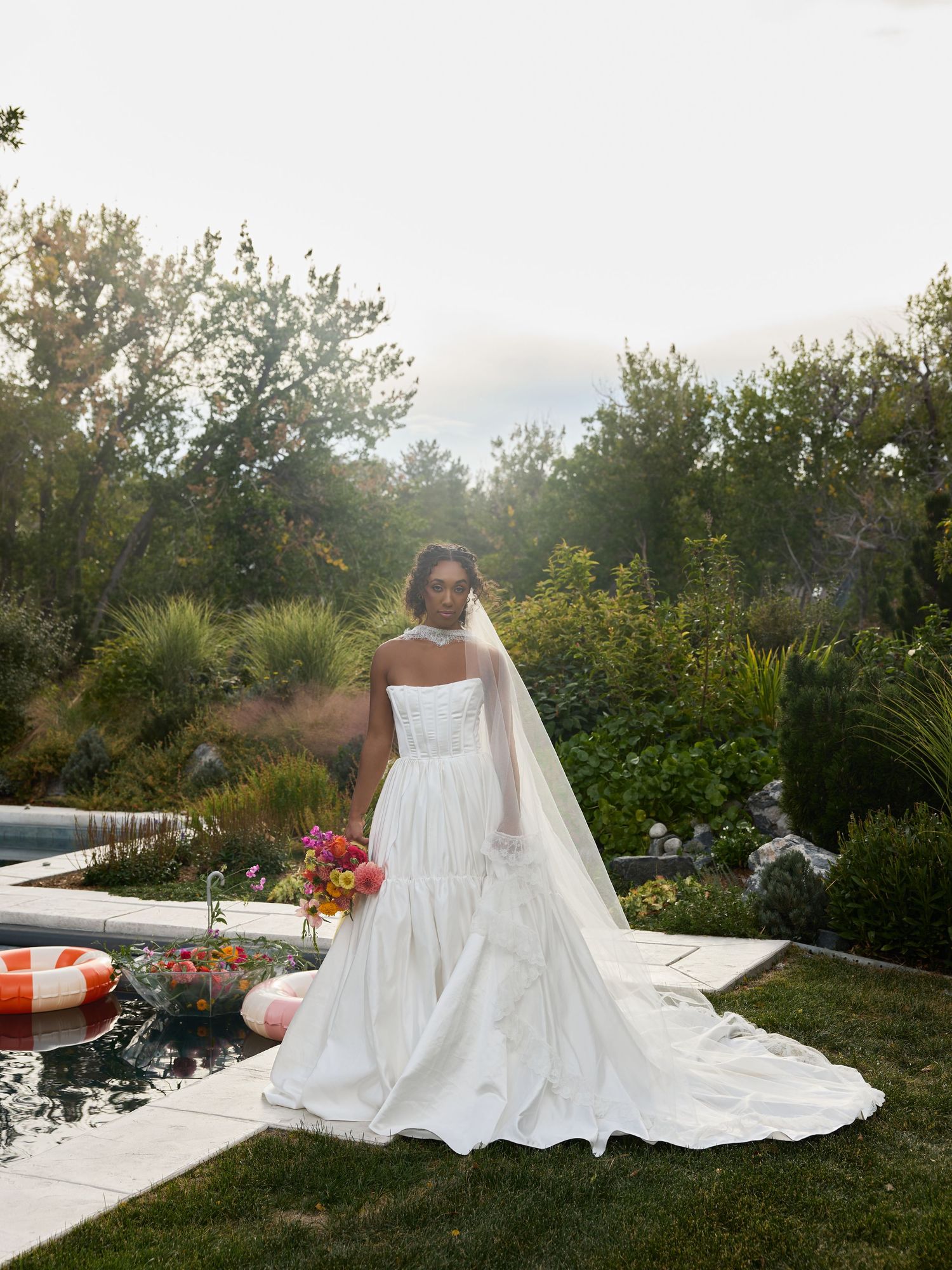 A bride in a strapless white gown and long veil holds a vibrant bouquet while standing beside a pool in a lush garden.