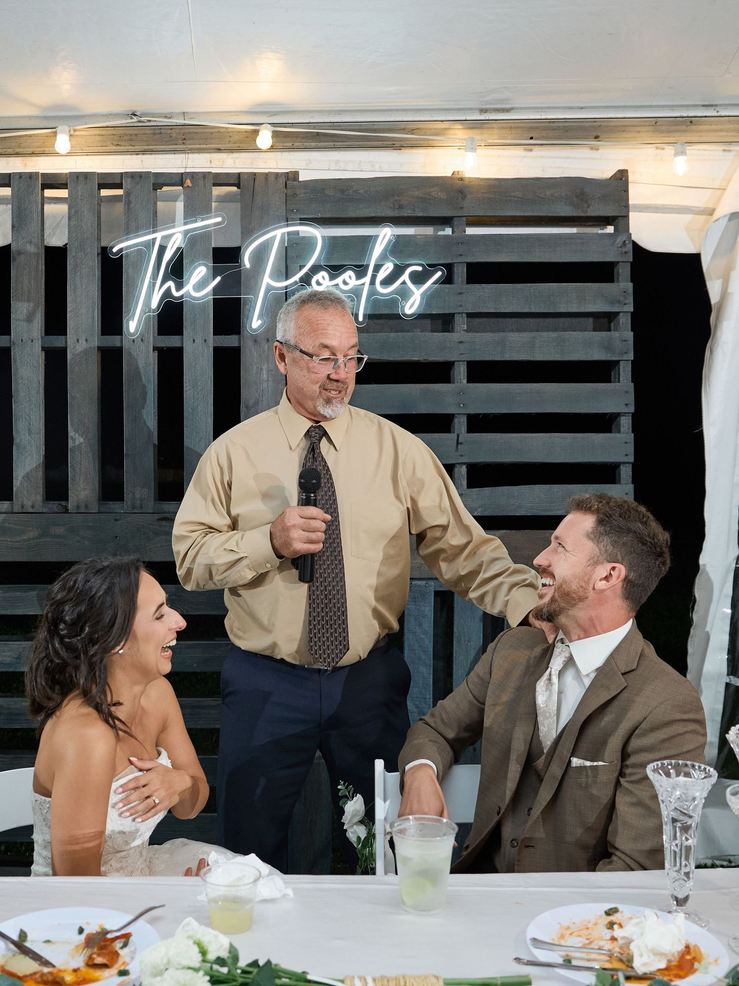 A man in a tie speaks into a microphone and rests a hand on the groom's shoulder as the smiling bride looks on at the reception.