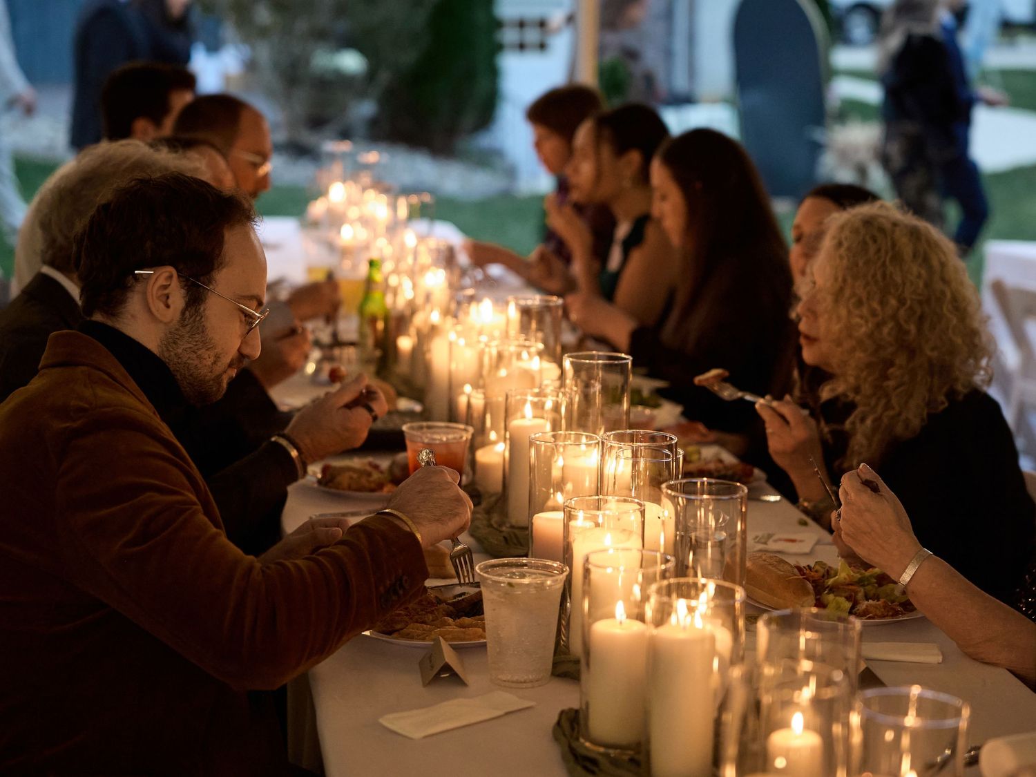 Guests dine together at a long candlelit table outdoors during a wedding dinner, with glowing candles lining the center.