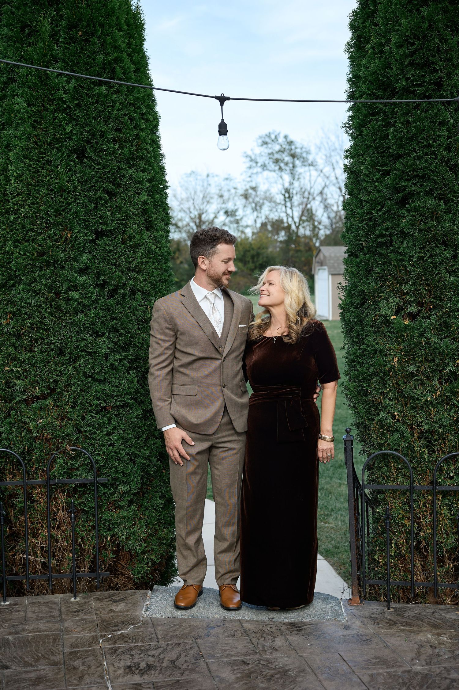 Groom in a tan suit and woman in a brown velvet dress embrace between tall evergreen hedges under string lights.
