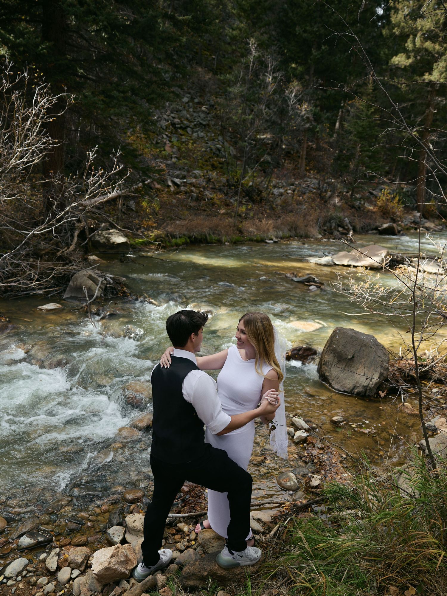 Bride and groom embrace on the rocky bank of a rushing mountain river during their outdoor elopement session.
