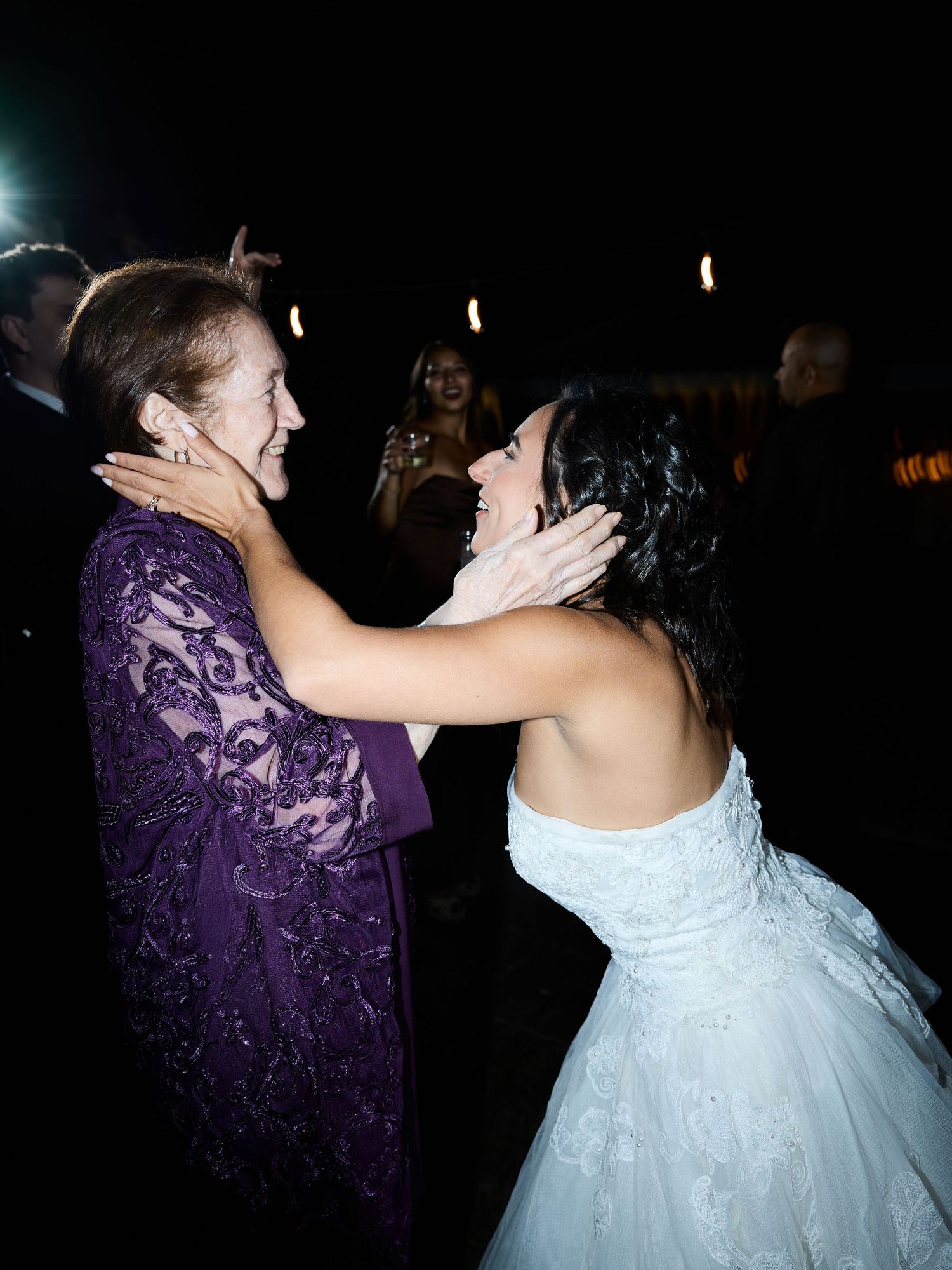 Bride in a strapless white gown shares a warm embrace with an older woman in a purple lace dress at the reception.