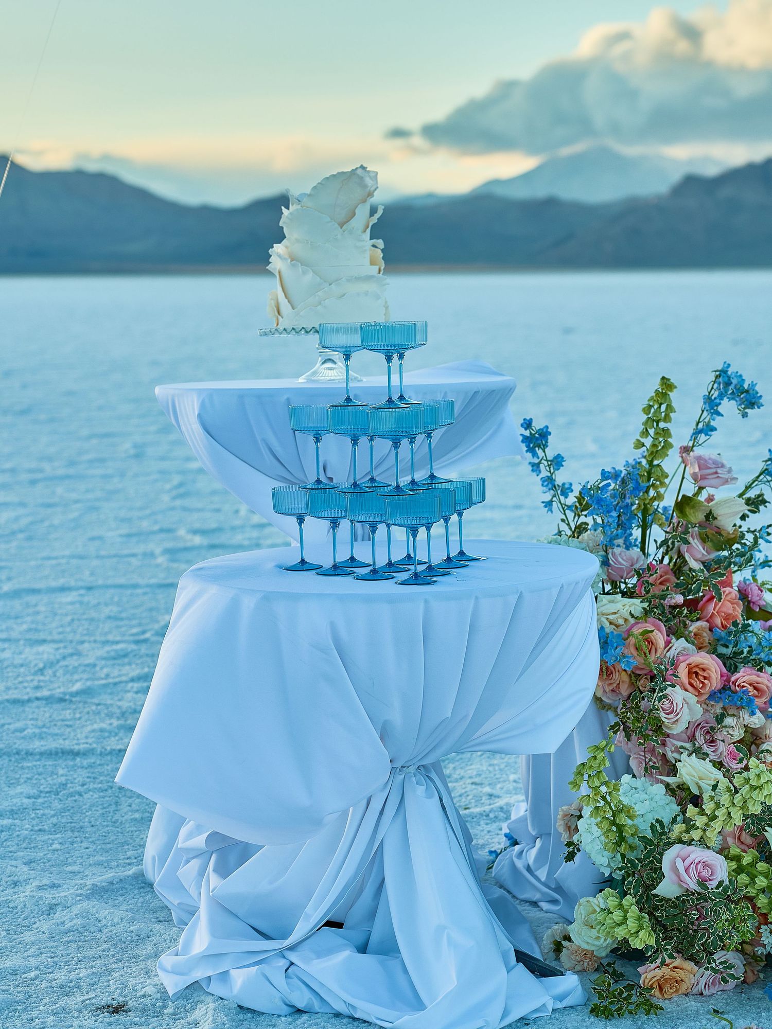 Tower of blue coupe glasses and a white sculpted wedding cake on a draped table beside a floral arrangement at a salt flat.