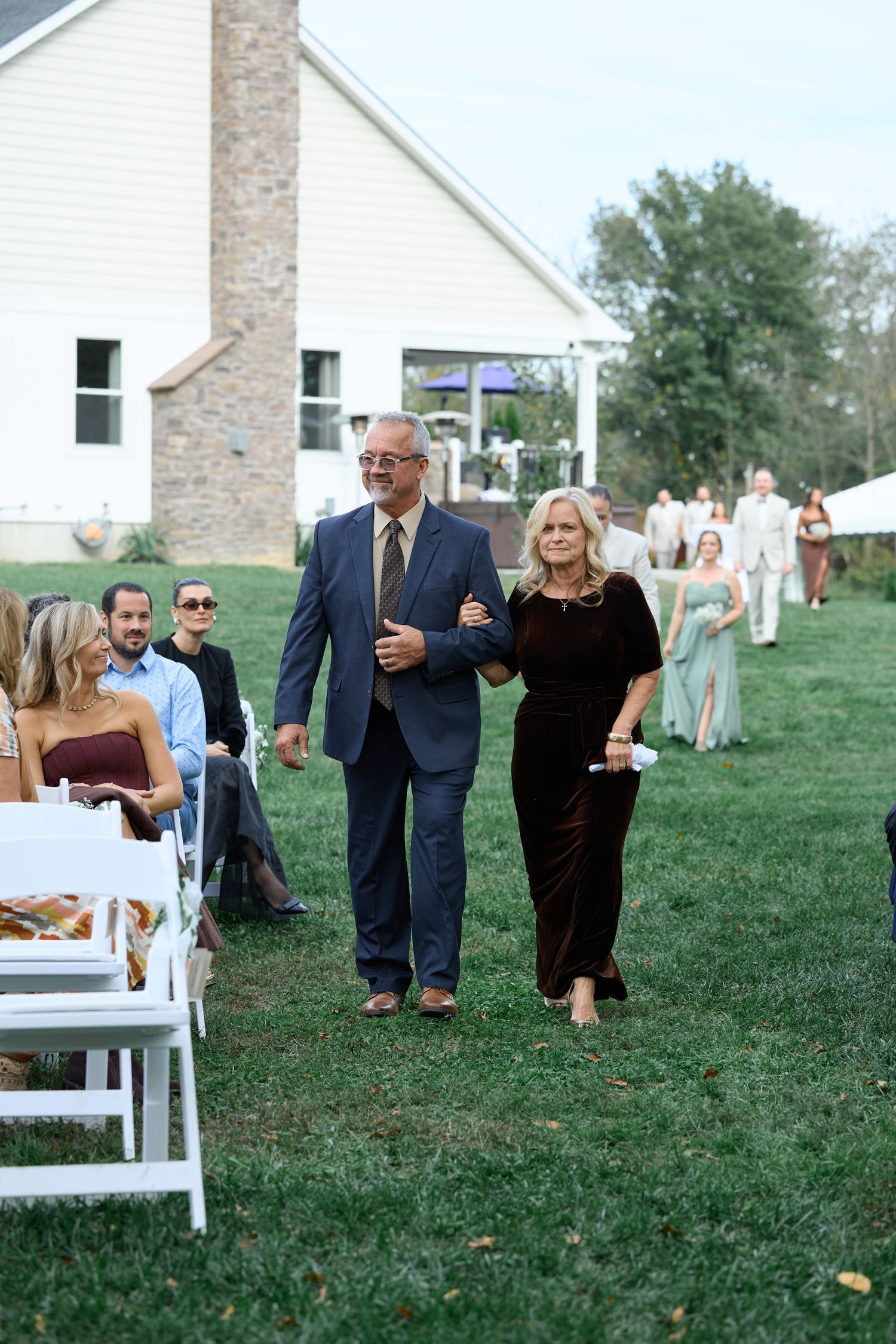 Smiling man in a navy suit walks arm in arm with a woman in a dark dress toward the outdoor wedding ceremony.