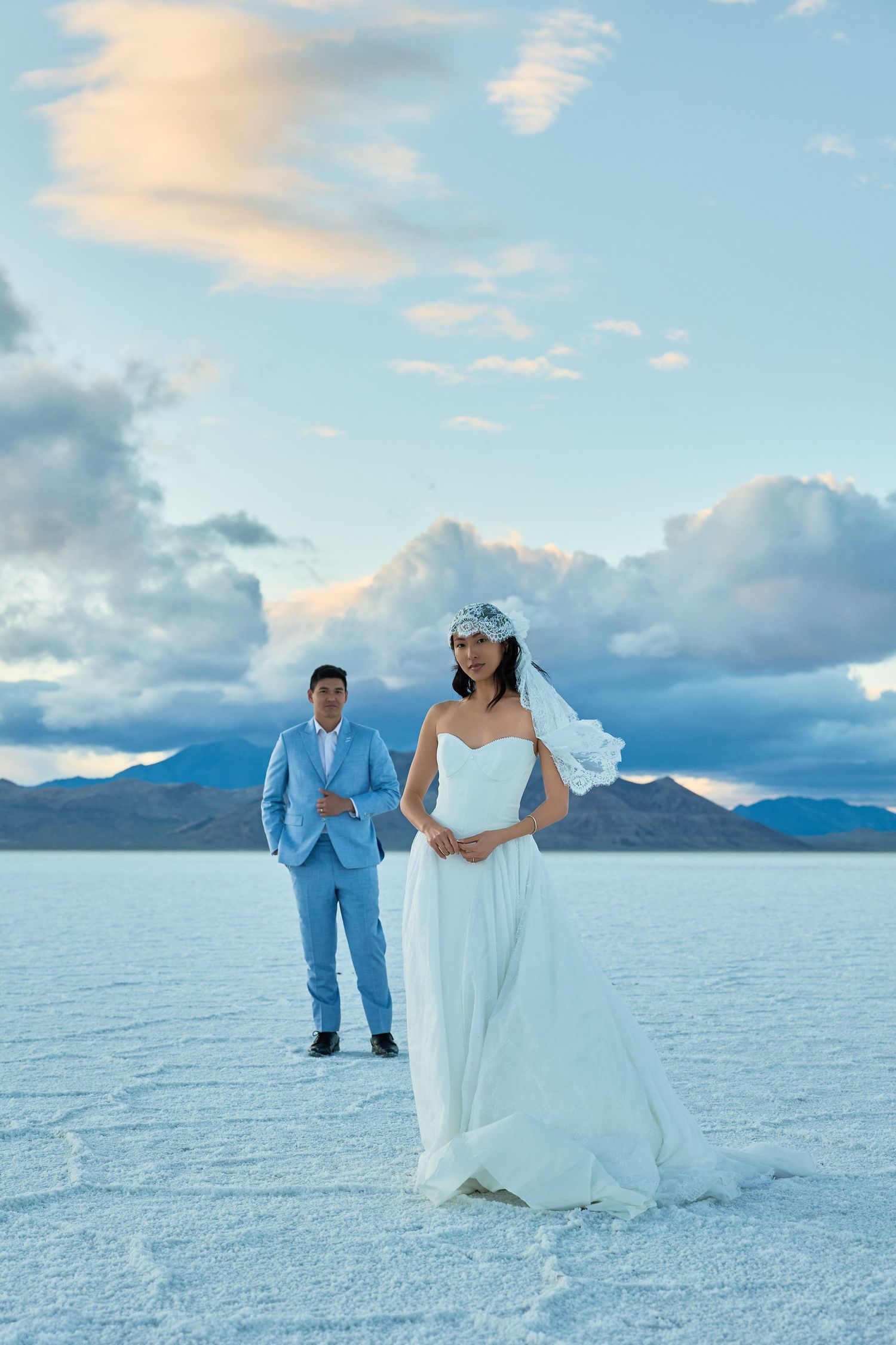 Groom in a light blue suit stands on a white salt flat while the bride in a white gown and veil poses in front under a cloudy sky.