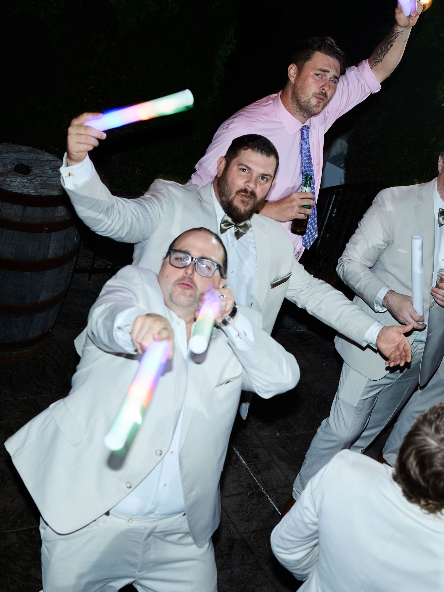 Groomsmen in cream suits pose playfully with glowing light sticks and drinks on the dance floor at a wedding reception.
