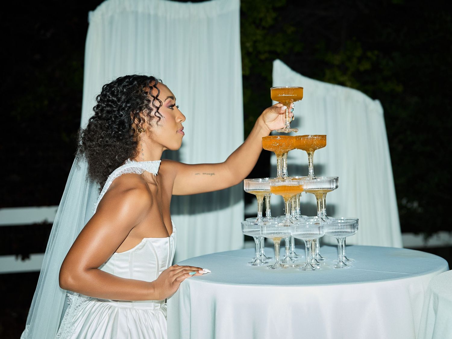 A bride in a white gown reaches up to take a coupe glass from a champagne tower set on a draped table at an evening wedding reception.